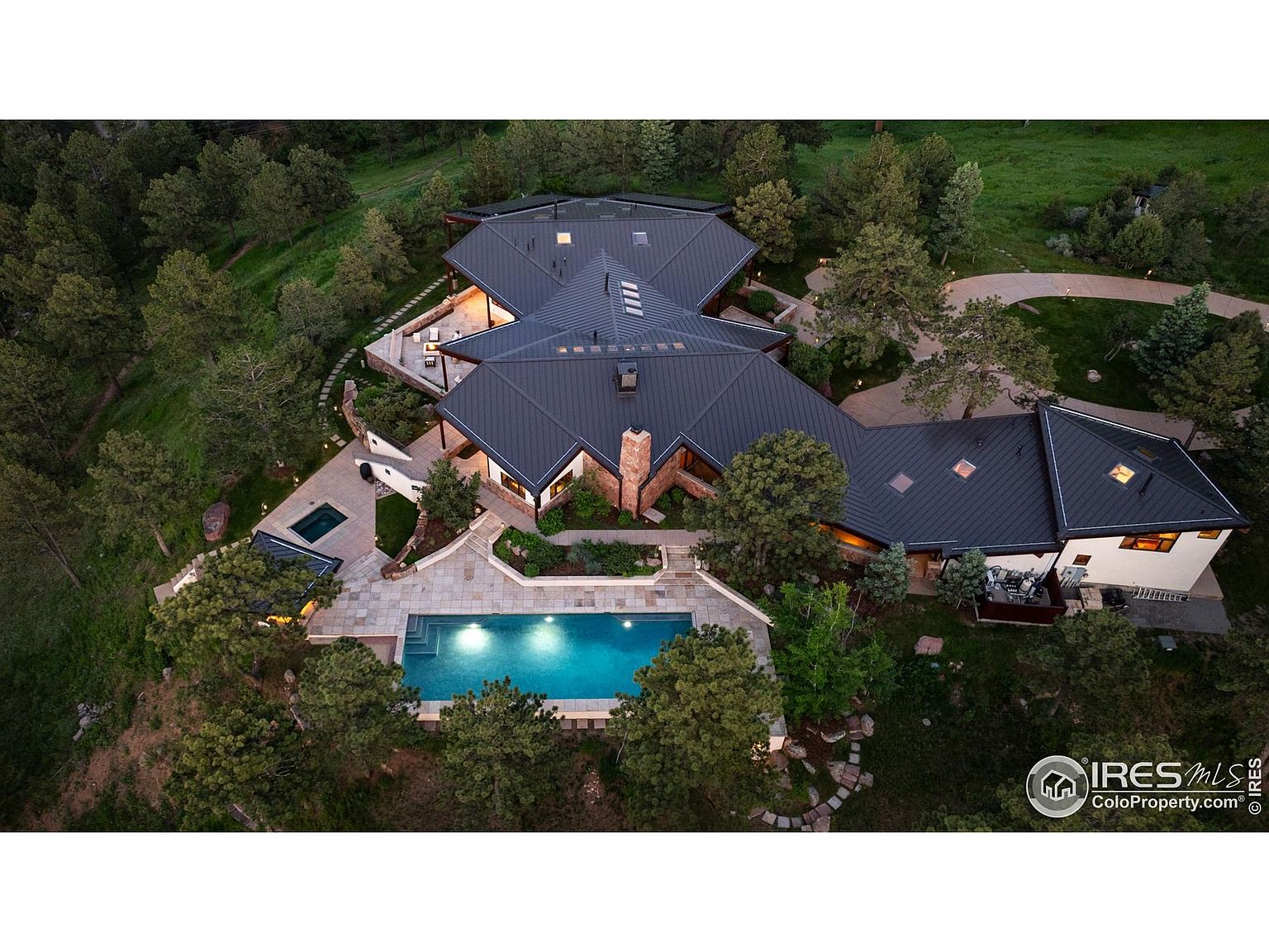 This aerial view showcases a luxurious estate nestled among lush greenery. The property features a unique architectural design with a dark roof, a sparkling blue swimming pool, and a separate spa area. The surrounding landscape includes mature trees and manicured gardens, creating a private and serene setting.