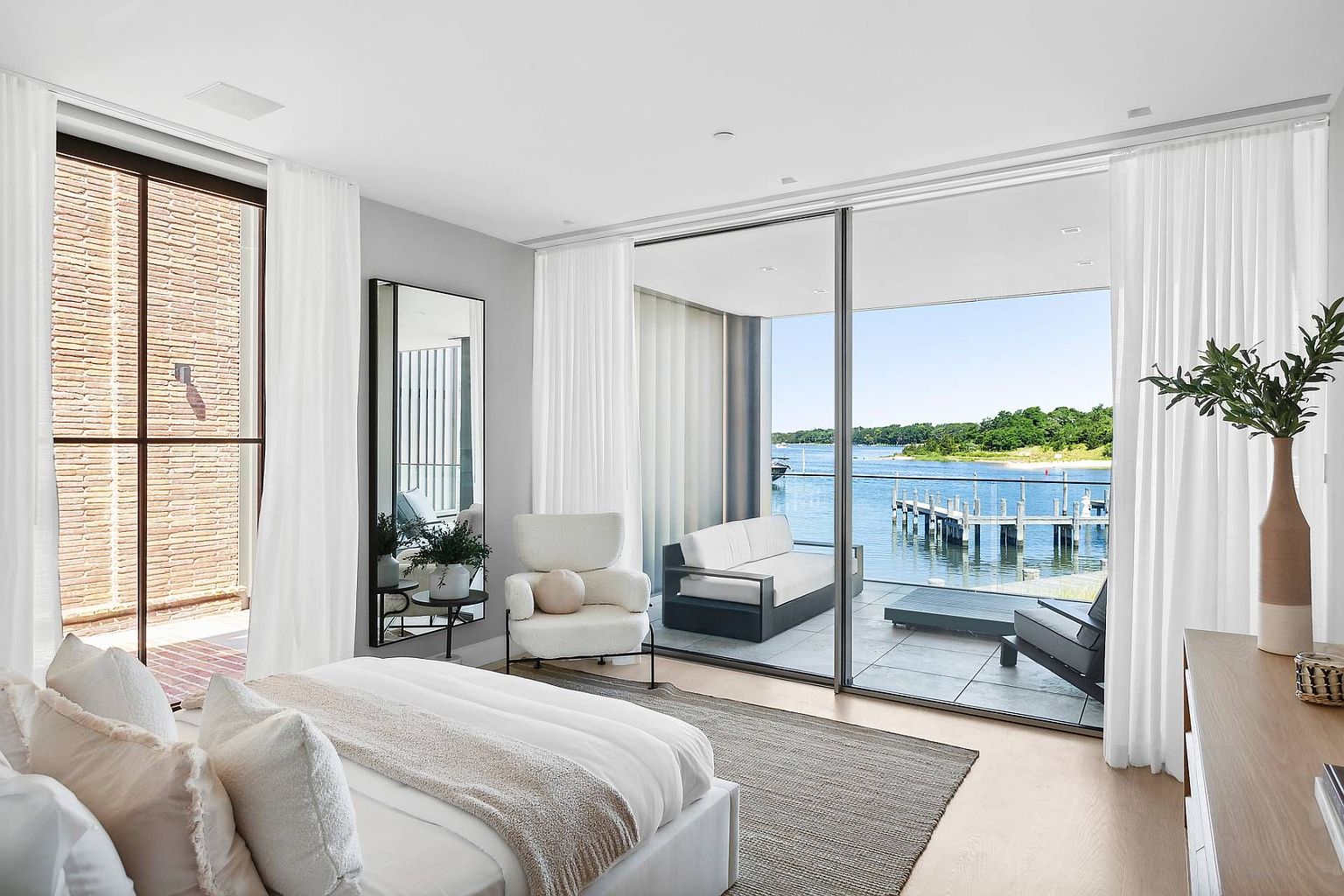 This serene primary bedroom features a neutral color palette and a seamless transition to a private balcony through expansive floor-to-ceiling glass sliding doors. The room is furnished with a plush bed, a textured area rug, and a cozy armchair, all oriented to maximize the stunning waterfront views. A tall, sleek mirror reflects the space, while the minimalist design emphasizes the connection between the indoor living area and the tranquil outdoor scenery.