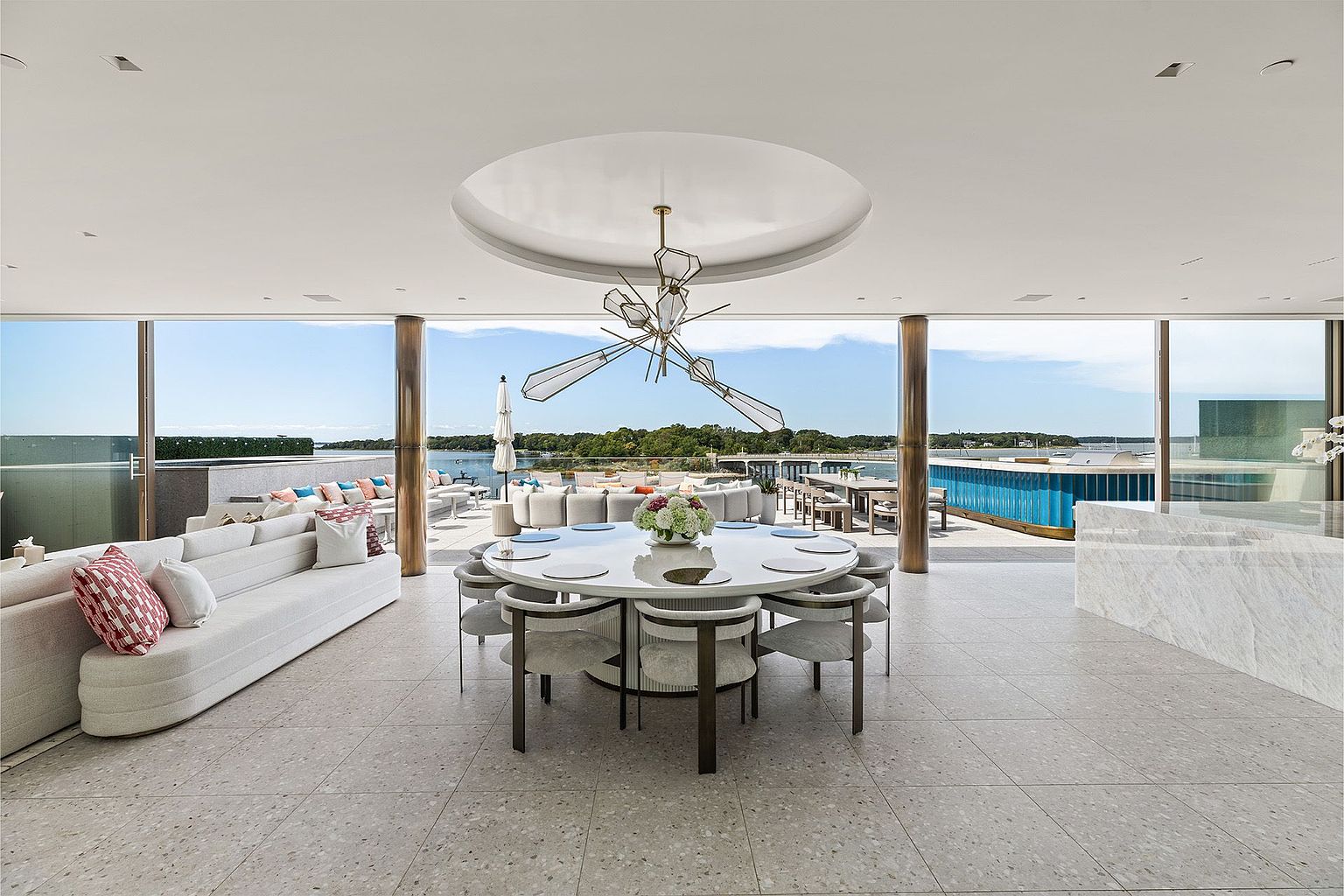 This expansive, open-concept dining area seamlessly transitions to a sprawling outdoor terrace through floor-to-ceiling glass walls. The space is anchored by a large, circular white dining table surrounded by modern, neutral-toned chairs, all positioned beneath a striking, sculptural geometric chandelier. The interior features light-toned stone flooring that extends onto the exterior deck, creating a cohesive indoor-outdoor living experience with panoramic water views.