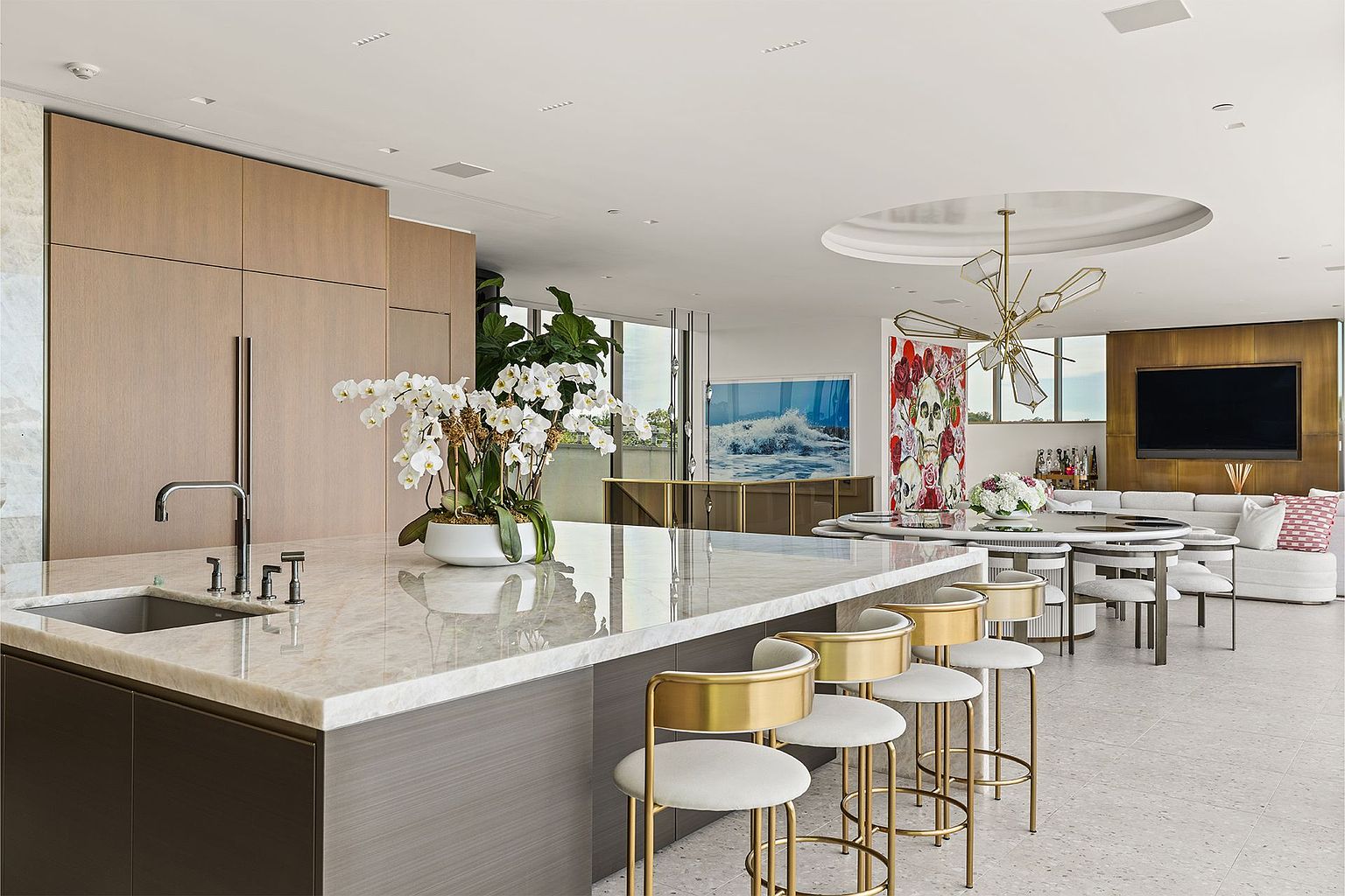 This sophisticated open-concept kitchen features a large stone-topped island with gold-toned bar stools and a sleek, integrated sink. The space is characterized by light wood cabinetry, a striking geometric chandelier, and a seamless transition into a dining and living area. The bright, airy atmosphere is enhanced by a large piece of vibrant wall art and a neutral color palette, creating a luxurious and modern living environment.