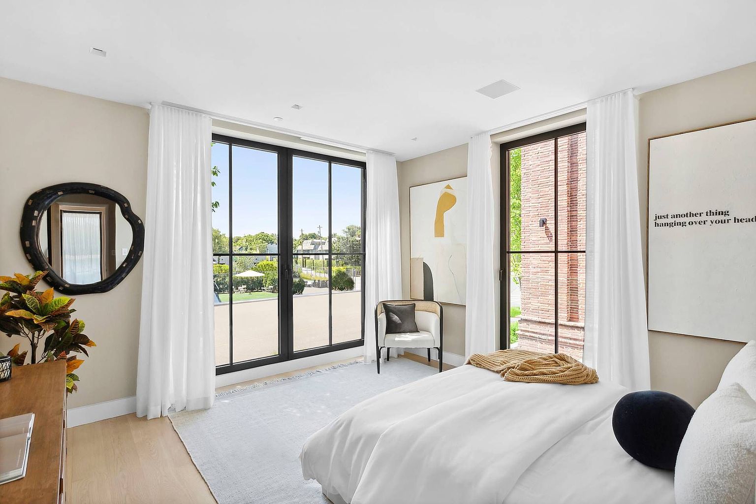 This bright and airy primary bedroom features a minimalist aesthetic with light wood flooring and neutral walls. Large windows with sheer white curtains allow for abundant natural light and provide a view of the outdoor patio area. The room is furnished with a comfortable bed, a modern accent chair, and unique wall art, creating a serene and sophisticated atmosphere.