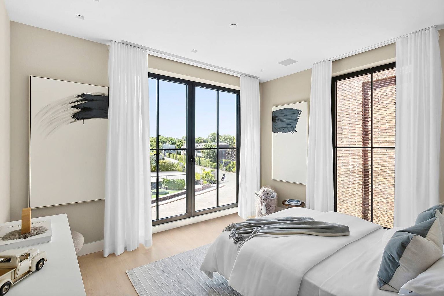 This bright and airy bedroom features a minimalist aesthetic with light wood flooring and neutral walls. The room is anchored by a bed dressed in white linens with grey accents, flanked by large windows that provide ample natural light and views of the surrounding neighborhood. Abstract art pieces hang on the walls, and a small decorative animal figure adds a touch of personality to the serene, modern space.