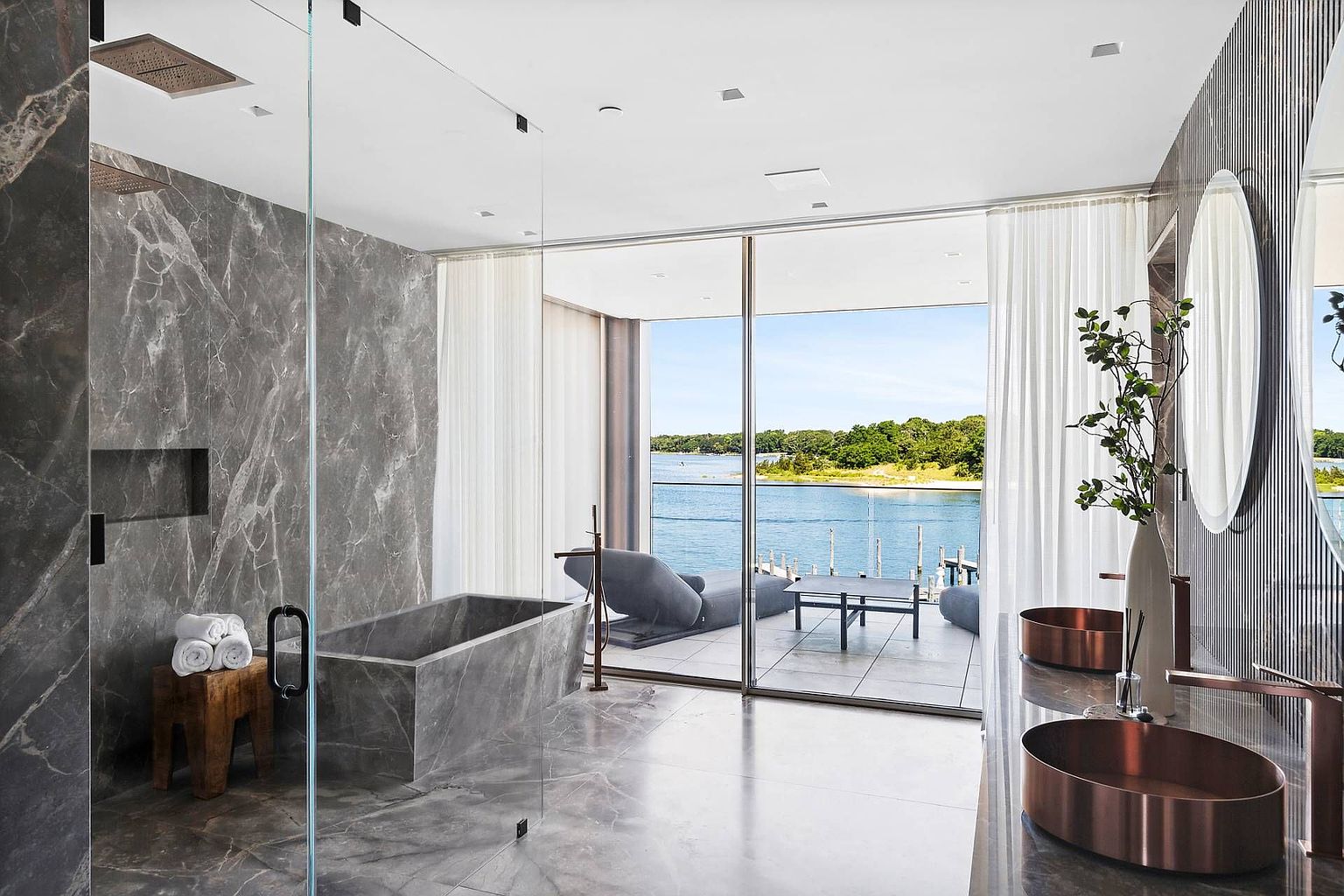 This luxurious primary bathroom features a striking stone-clad soaking tub and a glass-enclosed shower area, both set against dramatic, dark-veined stone walls. A pair of metallic vessel sinks sits atop a matching stone vanity, while floor-to-ceiling glass doors open directly onto a private balcony overlooking the water. The space exudes a sophisticated, spa-like atmosphere with a seamless transition between the indoor sanctuary and the scenic outdoor deck.