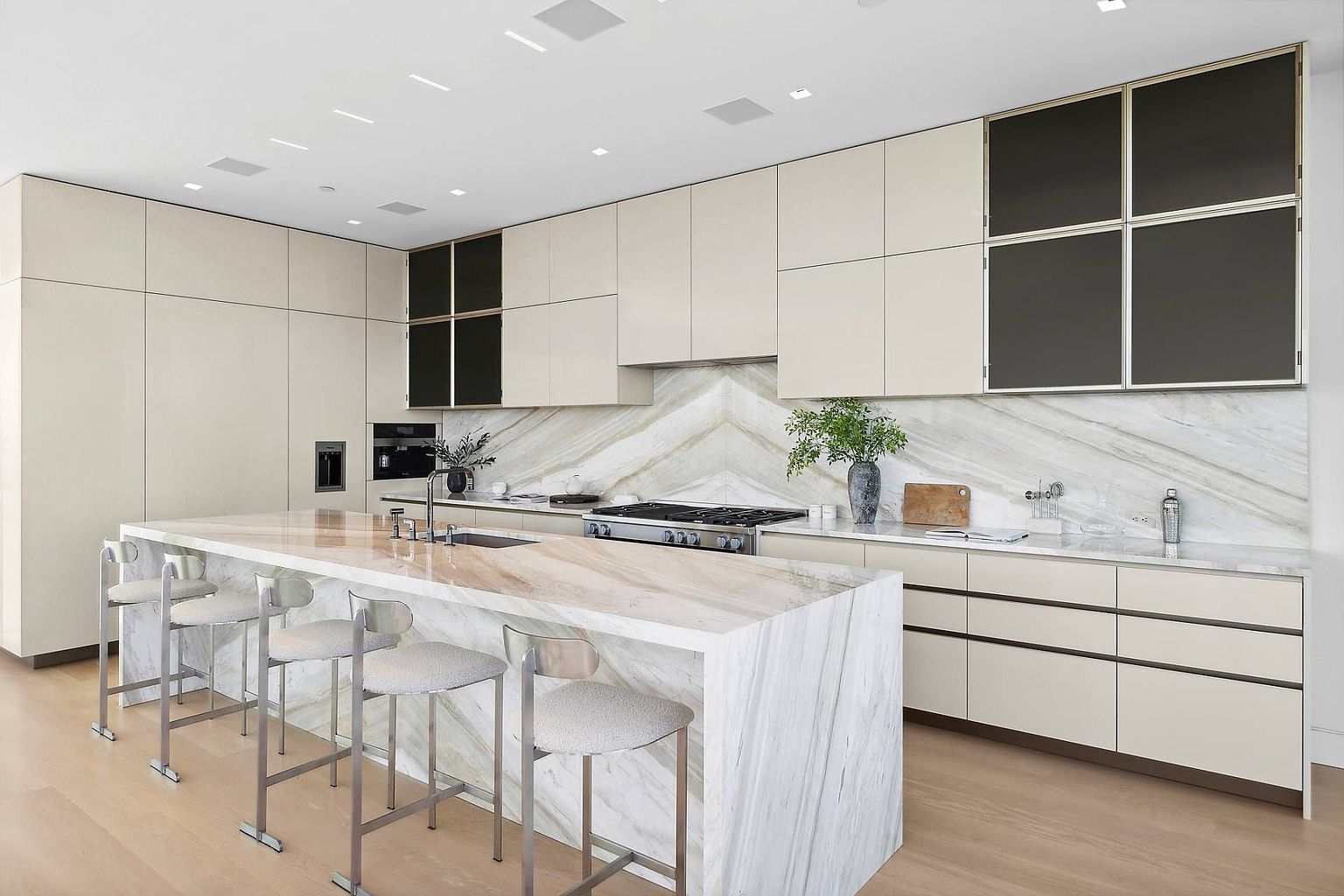 This sleek, contemporary kitchen features a large central island with a waterfall-edge stone countertop and four modern bar stools. The cabinetry is finished in a soft neutral tone, complemented by dark-framed glass-front upper cabinets and a striking chevron-patterned stone backsplash behind the gas range. The space is illuminated by recessed lighting and offers a clean, minimalist aesthetic with light-toned wood flooring.