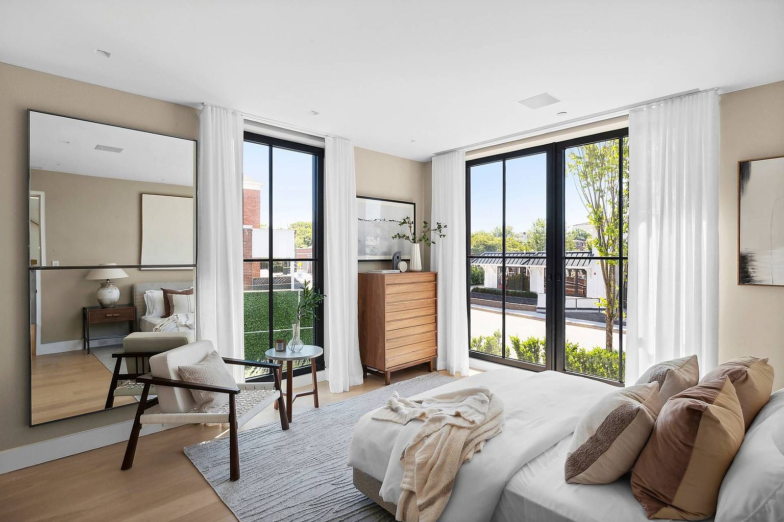 This bright and airy primary bedroom features a neutral color palette, light wood flooring, and a large area rug that anchors the space. The room is highlighted by two sets of tall windows with sheer curtains that allow for abundant natural light, while a large wall mirror reflects the bed and adds depth to the room. A wooden dresser and a modern accent chair provide functional elegance, creating a serene and sophisticated atmosphere.