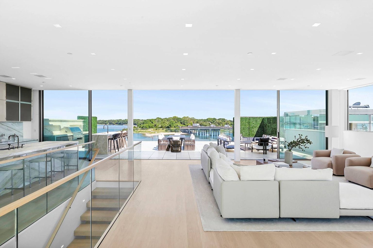 This expansive, open-concept living area features a bright, minimalist aesthetic with light wood flooring and a neutral color palette. The space is defined by massive floor-to-ceiling glass walls that seamlessly connect the interior to a spacious outdoor terrace overlooking a scenic waterfront and bridge. A large, plush sectional sofa anchors the living space, while a modern kitchen with a stone-topped island is visible to the left, creating a sophisticated and airy atmosphere.