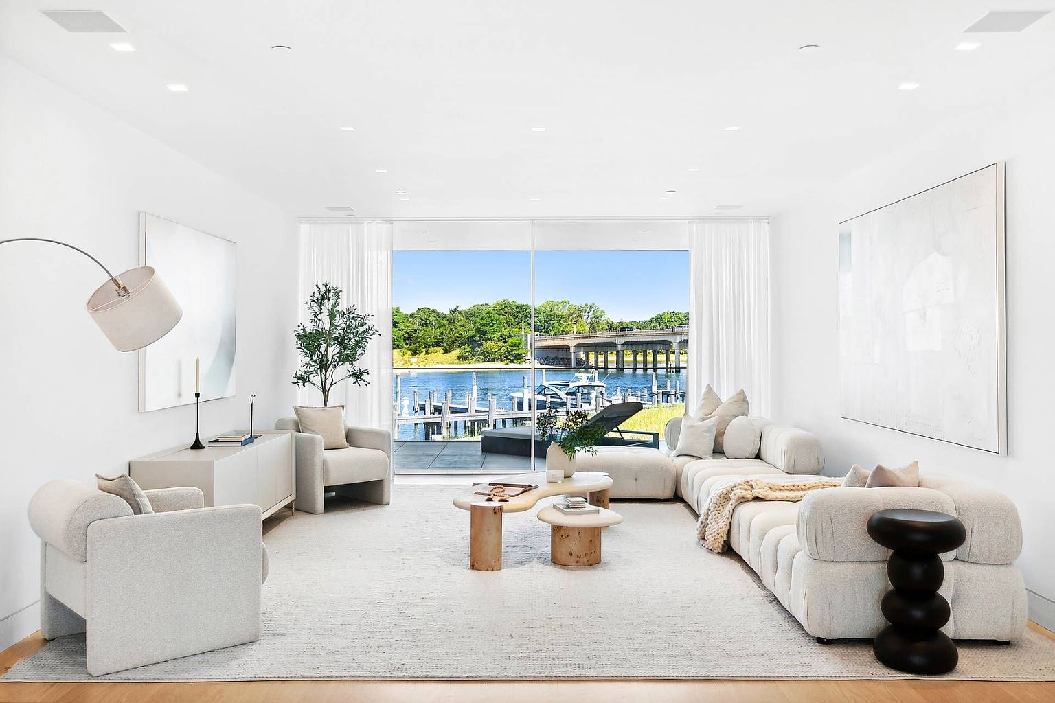 This bright and airy living room features a neutral color palette with a large, plush sectional sofa and matching armchairs arranged around a set of organic-shaped coffee tables. The space is anchored by a textured area rug and illuminated by a sleek, arched floor lamp and recessed ceiling lights. A wall of floor-to-ceiling glass sliding doors provides a seamless transition to an outdoor deck, offering a picturesque view of the water and a bridge in the distance.