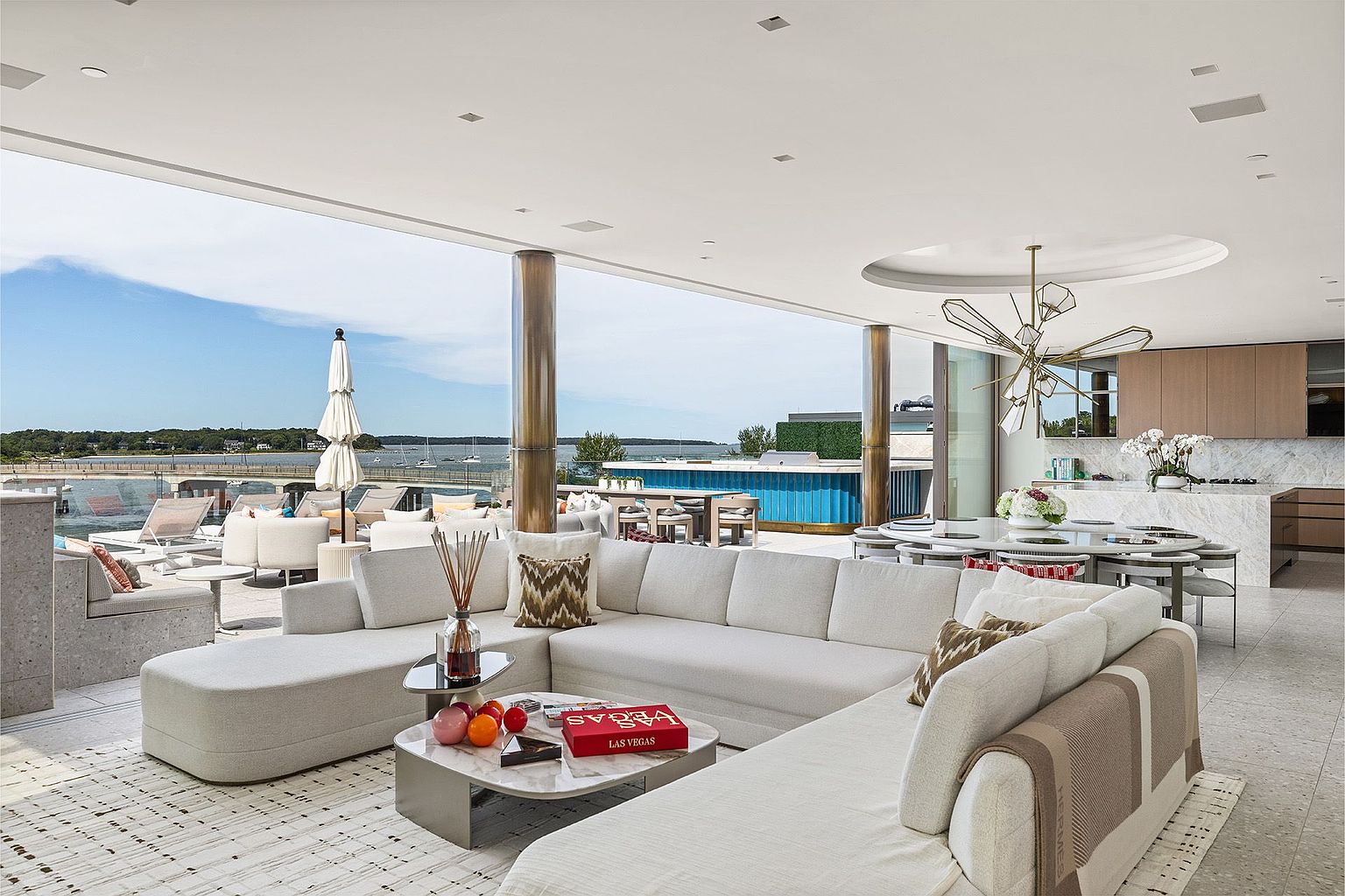 This expansive, open-concept living space seamlessly blends indoor and outdoor living through massive glass walls that open onto a sprawling terrace overlooking the water. The interior features a large, neutral-toned sectional sofa arranged around a central stone-topped coffee table, complemented by a sophisticated geometric chandelier above the adjacent dining area. The design emphasizes a luxurious, airy aesthetic with high-end finishes, light-toned flooring, and a panoramic view of the harbor and bridge.