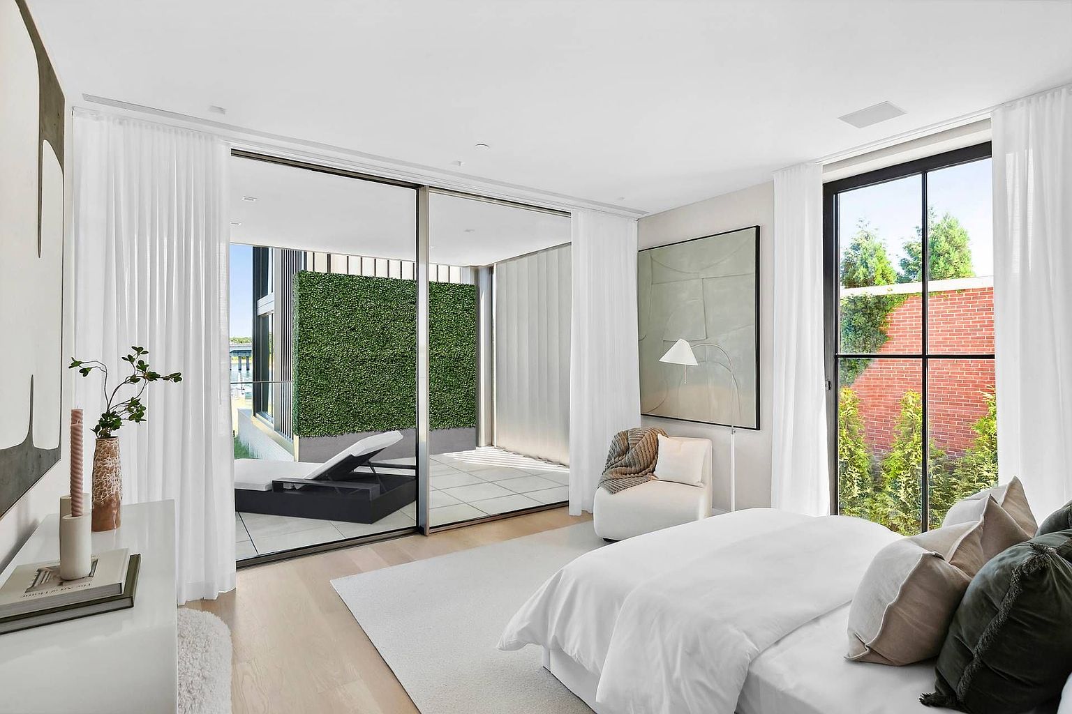 This bright and airy primary bedroom features a minimalist aesthetic with a neutral color palette and light wood flooring. A large sliding glass door provides seamless access to a private balcony, which is accented by a lush green privacy wall and a lounge chair. The room is furnished with a plush bed, a modern armchair, and a large abstract piece of art, creating a serene and sophisticated atmosphere.