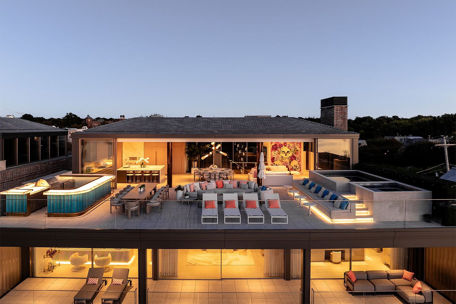This stunning aerial view captures a luxurious multi-level rooftop terrace designed for high-end entertaining. The upper deck features a vibrant blue-tiled bar, a long dining table, a lounge area with plush seating, and a raised spa, all illuminated by warm integrated lighting. The open-concept indoor living space seamlessly transitions to the outdoors through expansive glass walls, showcasing a modern aesthetic with sophisticated decor and a striking piece of wall art.