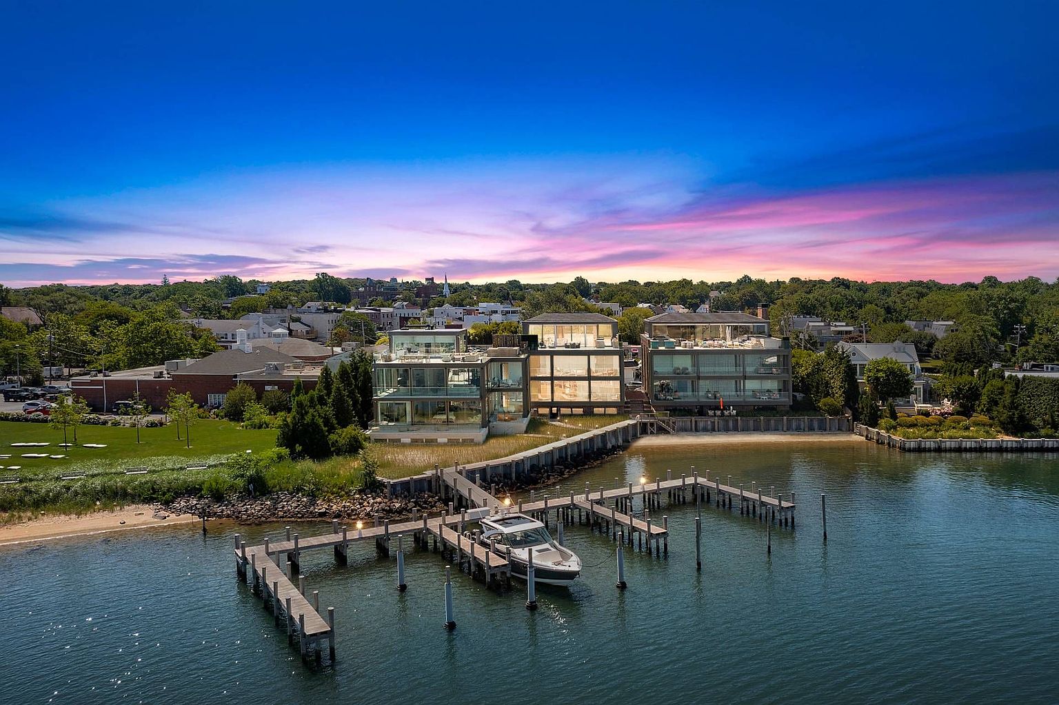 This stunning aerial view captures a modern, multi-level waterfront estate featuring expansive glass facades and multiple terraces overlooking the water. A private wooden dock extends into the calm bay, where a sleek motorboat is moored, emphasizing the property's luxury lifestyle and direct water access. The scene is set against a vibrant sunset sky, highlighting the architectural sophistication of the buildings and the surrounding lush coastal landscape.