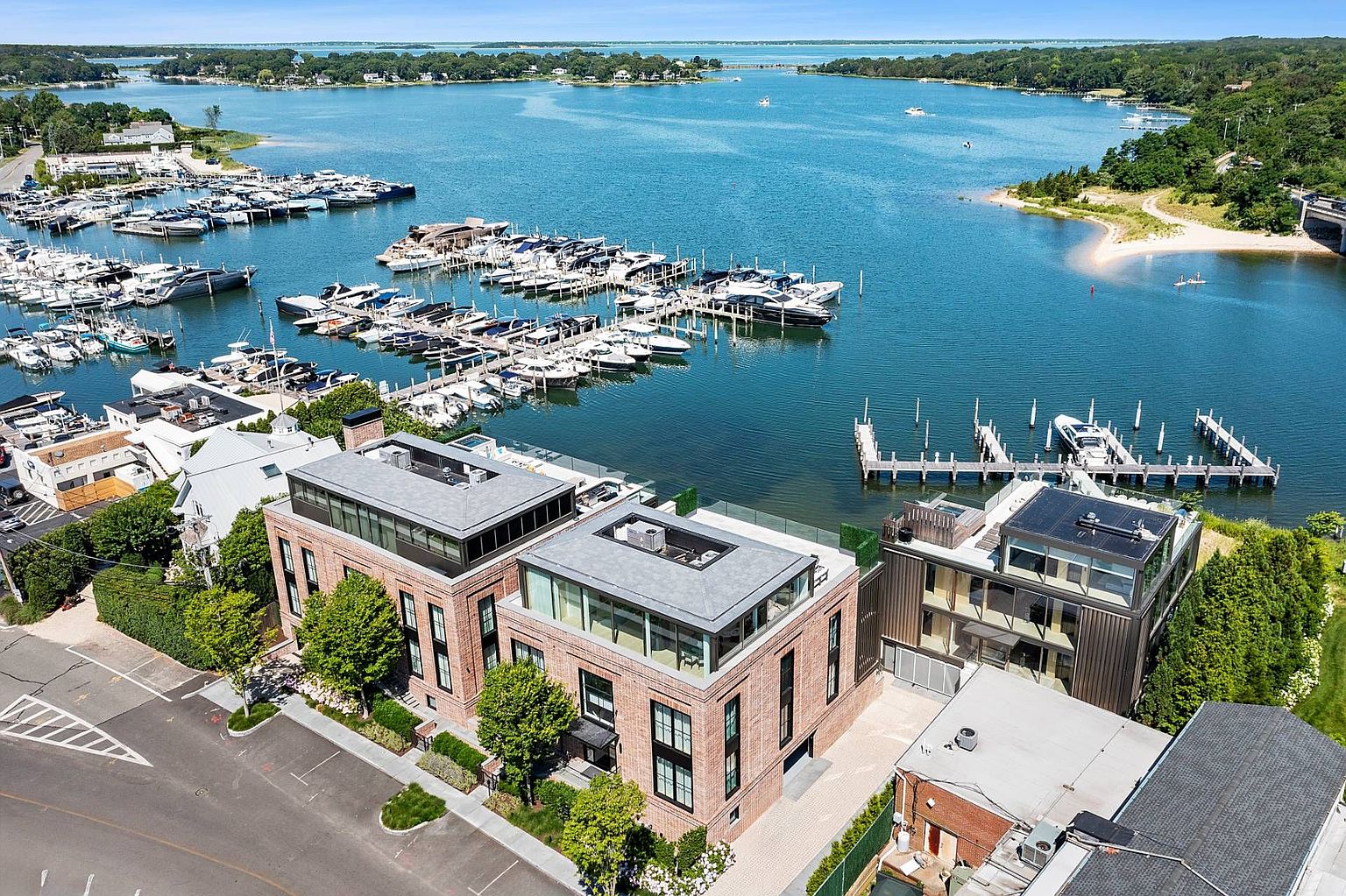 This high-angle aerial view captures a modern, multi-story brick residential building situated directly adjacent to a bustling marina. The property features expansive glass-enclosed rooftop levels and a sleek, contemporary design that contrasts with the surrounding waterfront landscape. The scene highlights the prime location, showcasing the proximity to numerous boat slips, a sandy shoreline, and the open water beyond.