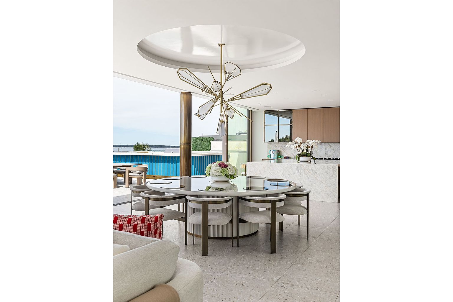This sophisticated dining area features a large, circular stone-topped table surrounded by curved, upholstered chairs, positioned beneath a striking, multi-armed geometric chandelier. The space flows seamlessly into an open-concept kitchen with light-toned cabinetry and a stone island, while large glass doors provide an expansive view of an outdoor deck and the water beyond. The interior design is characterized by a neutral color palette, clean lines, and a luxurious, contemporary aesthetic.
