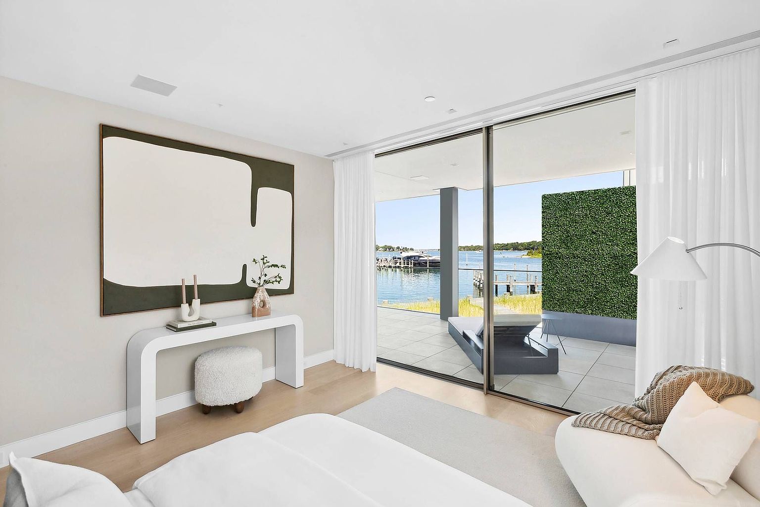 This bright and airy primary bedroom features a minimalist aesthetic with light wood flooring and a neutral color palette. A sleek white console table sits beneath a large abstract art piece, while floor-to-ceiling glass sliding doors provide a seamless transition to a private balcony overlooking the water. The room is furnished with a plush white sofa and a textured ottoman, creating a serene and luxurious atmosphere.