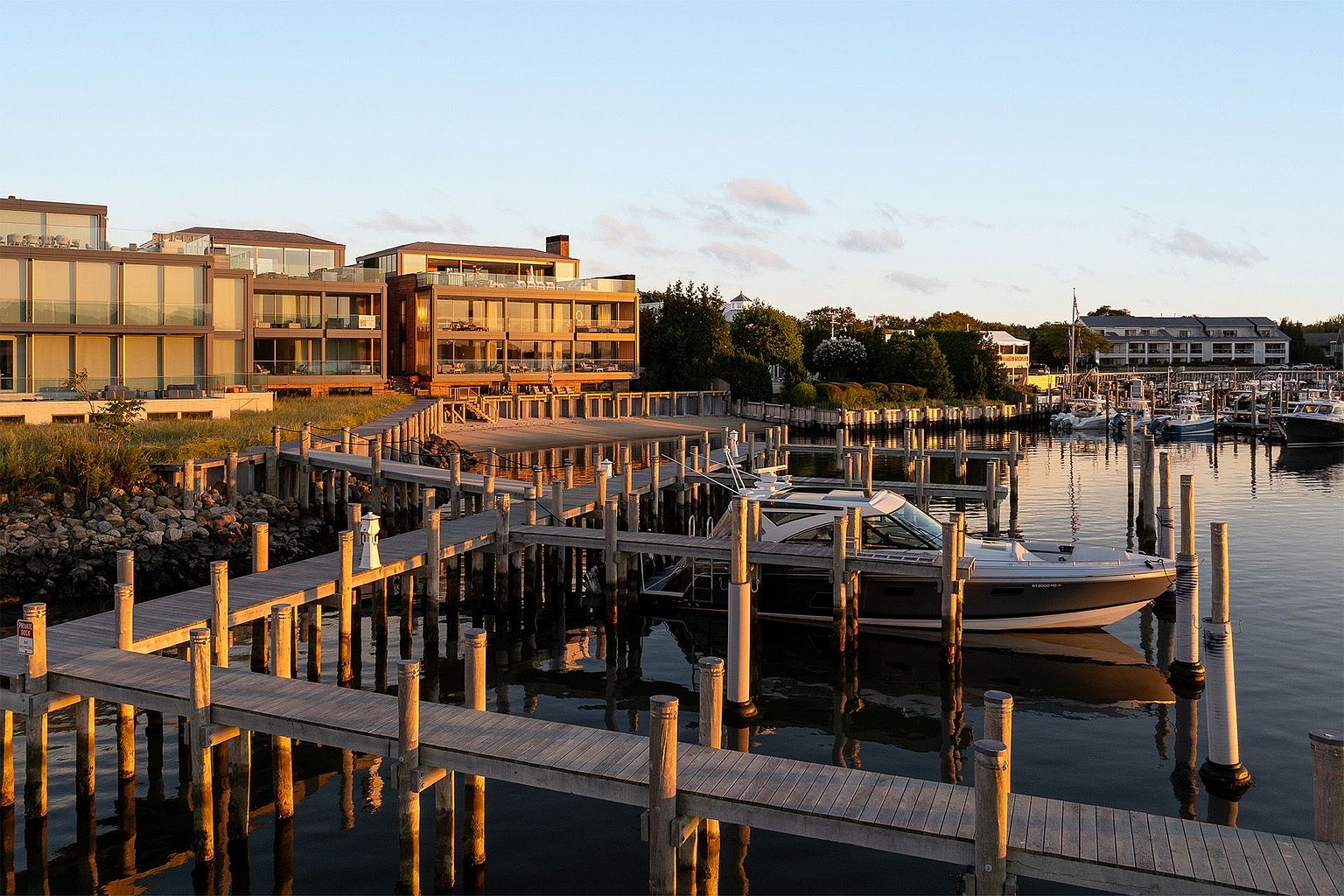 This scenic waterfront view captures a modern multi-story residence featuring extensive glass walls and expansive balconies overlooking a private dock and marina. The golden hour light casts a warm glow over the wooden pier, where a sleek motorboat is moored, creating a tranquil and luxurious atmosphere. The perspective is elevated, showcasing the seamless integration between the high-end architecture and the surrounding coastal landscape.