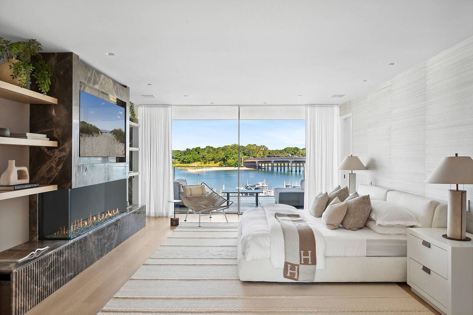 This luxurious primary bedroom features a minimalist aesthetic with a neutral color palette and a prominent bed adorned with a designer throw. A sleek, linear fireplace is set into a dark stone-clad chimney breast that extends upward, flanked by open shelving and a wall-mounted television. Large glass panels provide an unobstructed view of the waterfront, bridge, and outdoor deck, creating a seamless connection between the interior space and the scenic exterior.