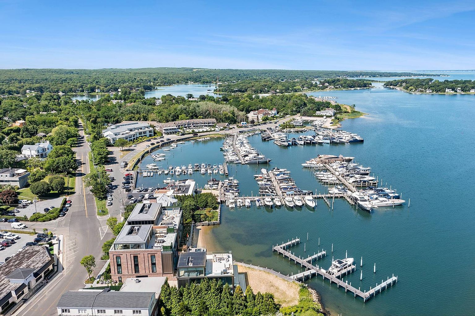 This high-angle aerial view captures a vibrant coastal marina filled with numerous boats docked in calm, blue waters. The scene includes a mix of modern multi-story residential buildings, a winding coastal road, and lush green landscapes surrounding the harbor. The perspective provides a comprehensive look at the waterfront lifestyle and the proximity of the residential properties to the boating facilities.