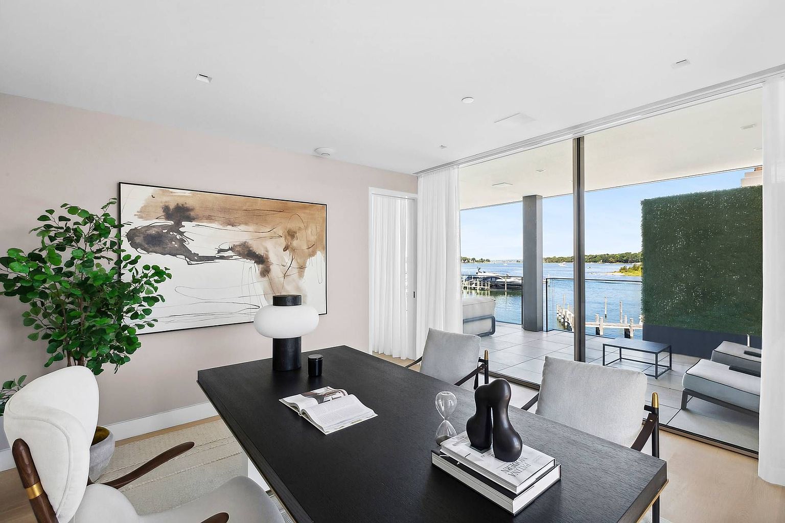 This sophisticated home office features a dark wood desk as the centerpiece, complemented by modern upholstered chairs and a minimalist table lamp. A large abstract painting hangs on the soft-toned wall, while floor-to-ceiling glass doors provide a seamless transition to a private balcony overlooking the water. The space exudes a serene, professional atmosphere with a clear focus on the scenic outdoor view.