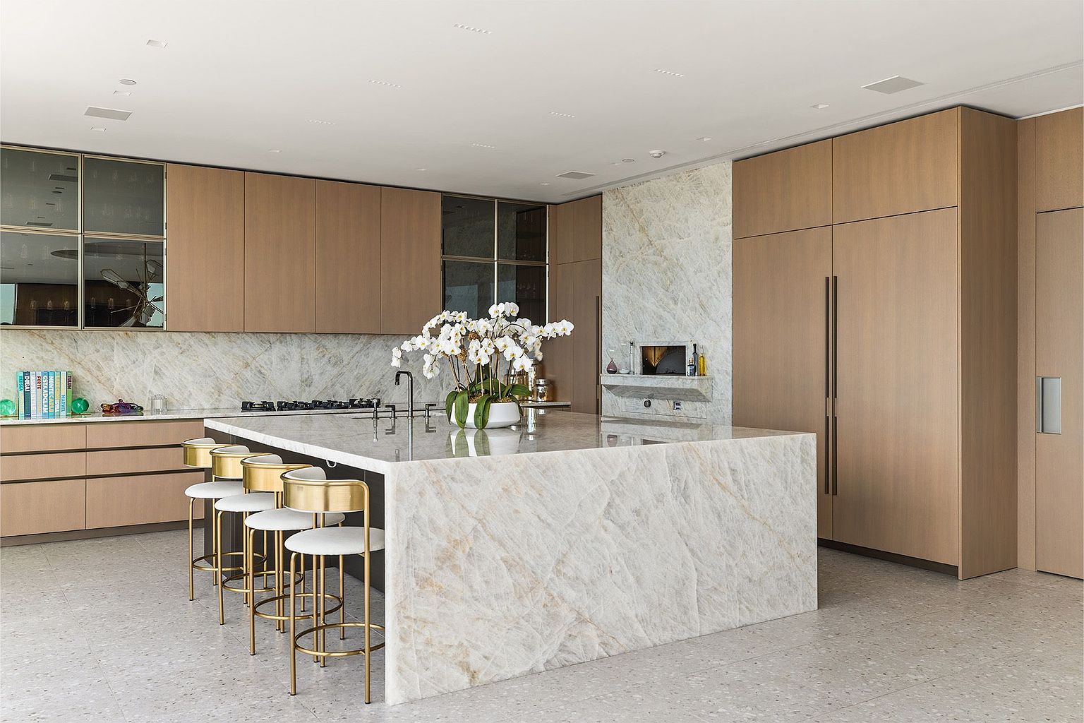 This sophisticated, modern kitchen features a large central island with a waterfall-edge stone countertop and gold-toned bar stools. The space is defined by light wood cabinetry, a stone-clad chimney breast, and a neutral color palette that creates a clean, high-end aesthetic. The perspective is a wide-angle shot capturing the seamless integration of the island, cabinetry, and open-concept layout.