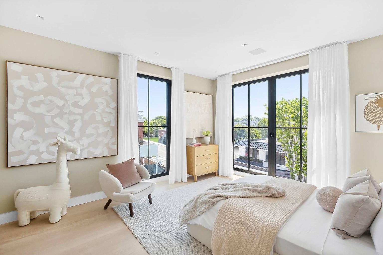 This bright and airy primary bedroom features a neutral color palette with soft textures and modern decor. The room is anchored by a bed with plush bedding and is illuminated by large windows that allow for abundant natural light. A whimsical llama-shaped accent piece and abstract wall art add a playful yet sophisticated touch to the serene space.
