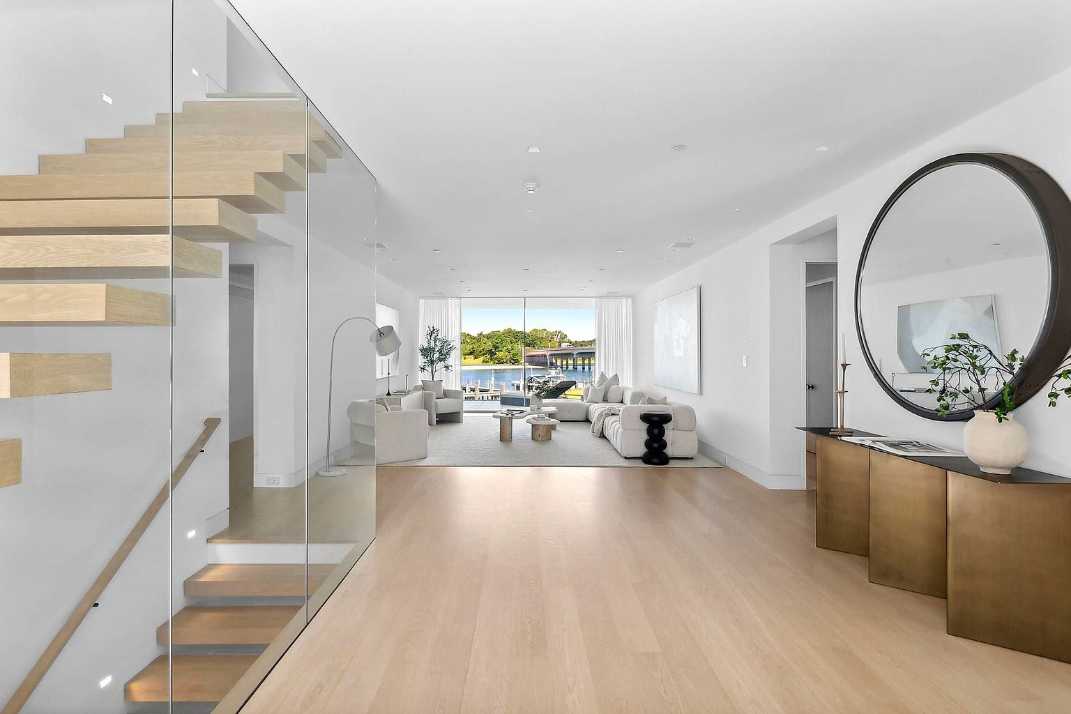 This bright, open-concept living space features light wood flooring and a minimalist aesthetic that leads the eye toward a large glass wall overlooking a waterfront view. To the left, a floating staircase with a glass railing adds a modern architectural element, while a large circular mirror and a metallic console table anchor the entryway on the right. The room is furnished with neutral-toned seating, creating a serene and sophisticated atmosphere perfect for luxury living.
