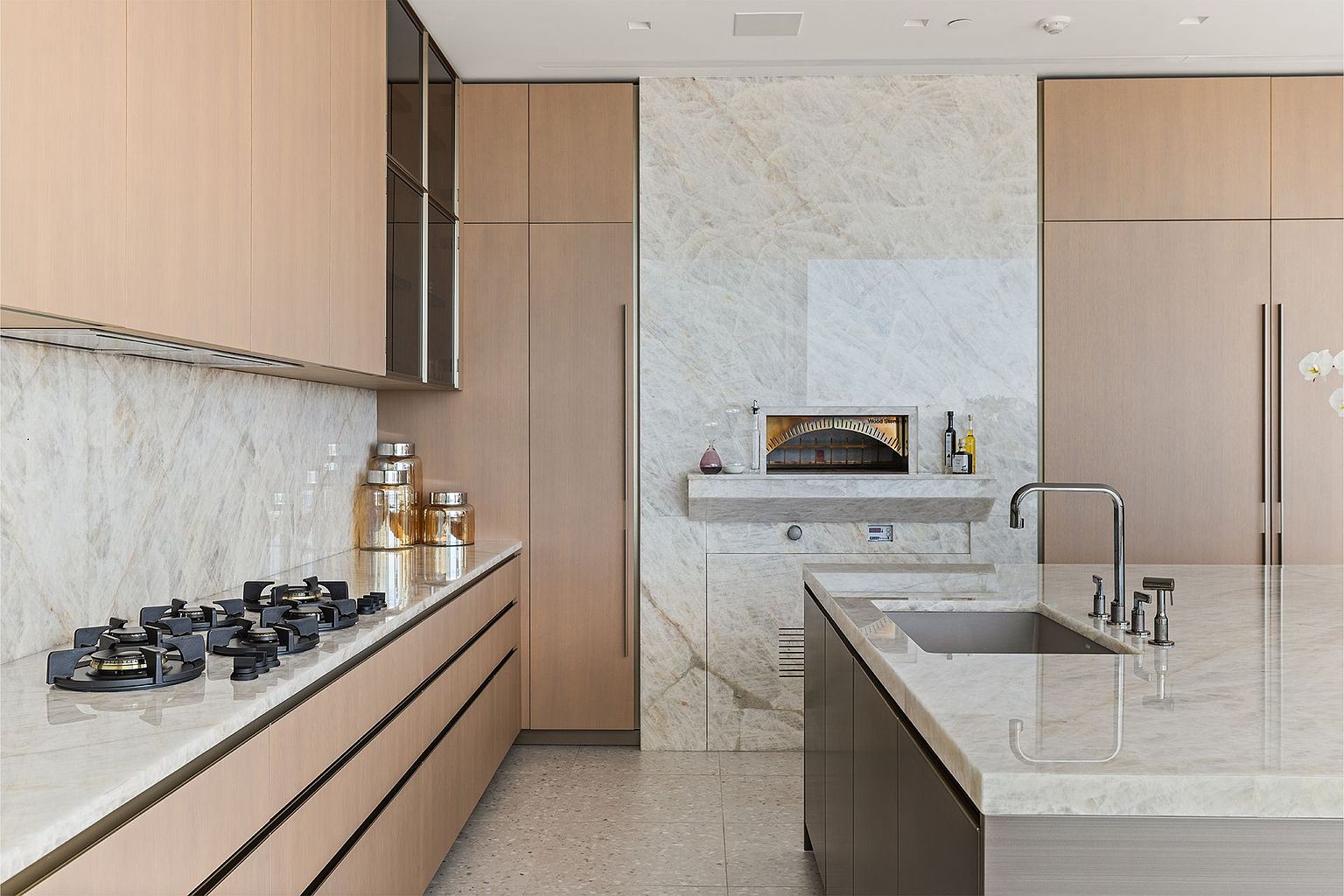 This sophisticated, modern kitchen features light wood cabinetry, a large stone-topped island with an integrated sink, and a gas cooktop set into a long stone counter. A standout feature is the built-in stone-clad oven set into a tall, light-toned stone wall, creating a seamless and luxurious aesthetic. The perspective is a wide-angle shot from the kitchen island, showcasing the clean lines, high-end finishes, and the open, airy layout of the space.
