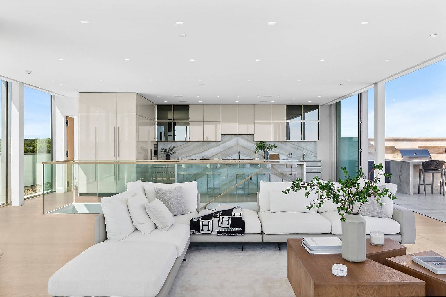 This open-concept living space features a large, plush white sectional sofa centered in the room, accented by a patterned throw blanket and a wooden coffee table. Beyond the seating area, a sleek kitchen with light-toned cabinetry and a stone backsplash is visible, separated by a glass-paneled railing. The room is bathed in natural light from expansive glass walls that offer views of the outdoors, creating a bright, airy, and contemporary atmosphere.