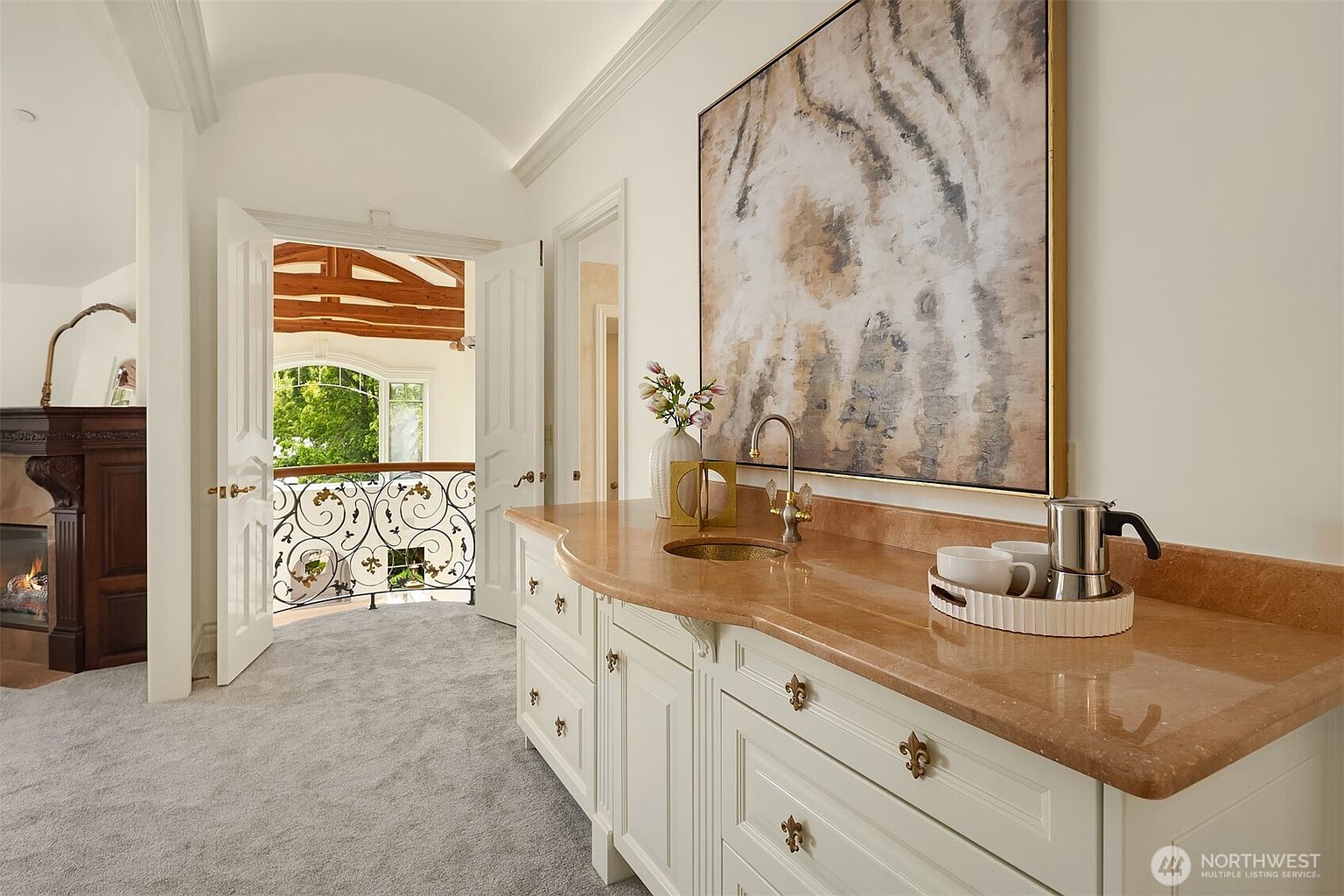 This elegant hallway features a built-in wet bar with a polished stone countertop, a small sink, and ornate gold-toned hardware, set against a backdrop of a large, abstract tiger-themed painting. The space leads through double doors to a balcony with intricate wrought-iron railings, offering a view of the outdoors. The combination of classic cabinetry, soft carpet, and architectural detailing creates a sophisticated and luxurious atmosphere.