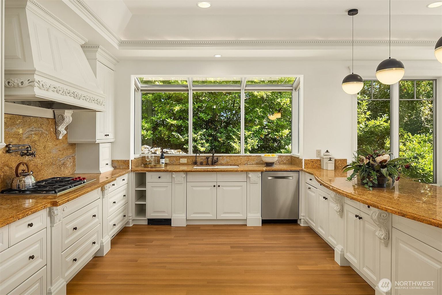 This elegant, U-shaped kitchen features classic white cabinetry with ornate corbel details, contrasted by warm, speckled granite countertops and rich hardwood flooring. A large, multi-pane window above the sink provides a bright, airy view of lush greenery, while modern spherical pendant lights hang over the peninsula. The space exudes a sophisticated, traditional charm with high-end finishes and a clean, well-organized layout.