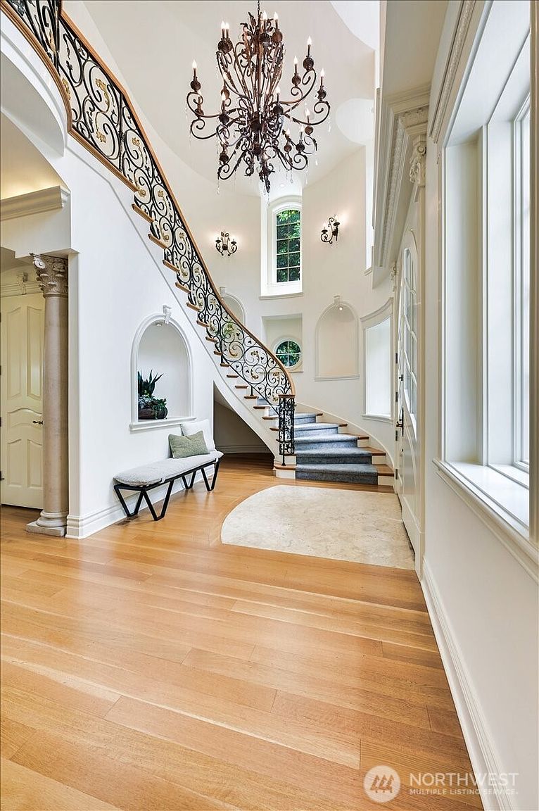 This grand foyer features a sweeping curved staircase with intricate wrought-iron railings and a dramatic, multi-tiered chandelier that serves as the focal point. The space is characterized by high ceilings, light hardwood flooring, and elegant architectural details like arched wall niches and a classic column. The bright, airy atmosphere creates a sophisticated and welcoming first impression for this luxury residence.