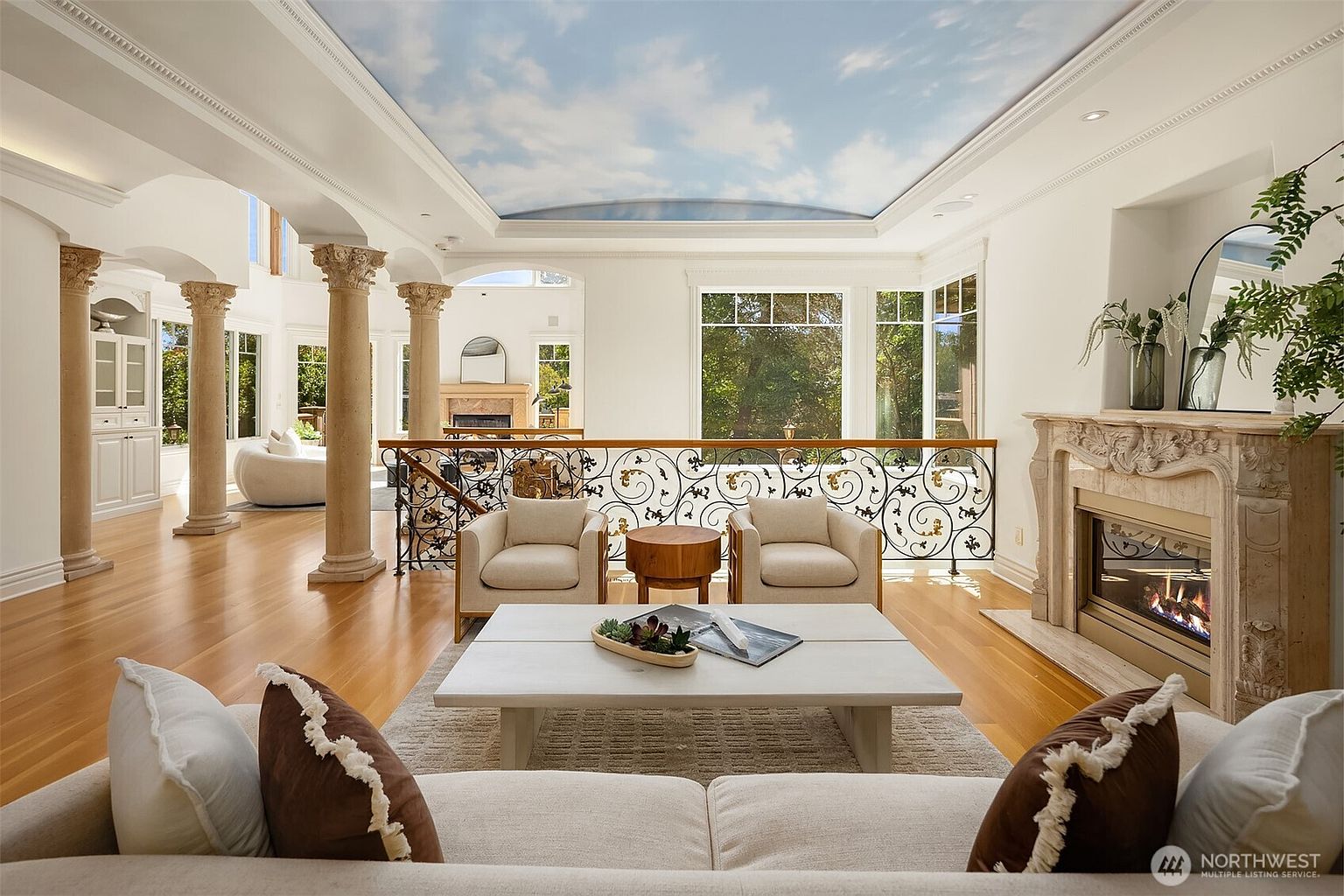 This elegant, expansive living room features a dramatic sky-painted ceiling and classic architectural columns that frame the space. The room is anchored by a sophisticated stone fireplace and a wrought-iron railing overlooking a lower level, creating a luxurious and airy atmosphere. Large windows flood the area with natural light, highlighting the polished hardwood floors and refined, neutral-toned furnishings.