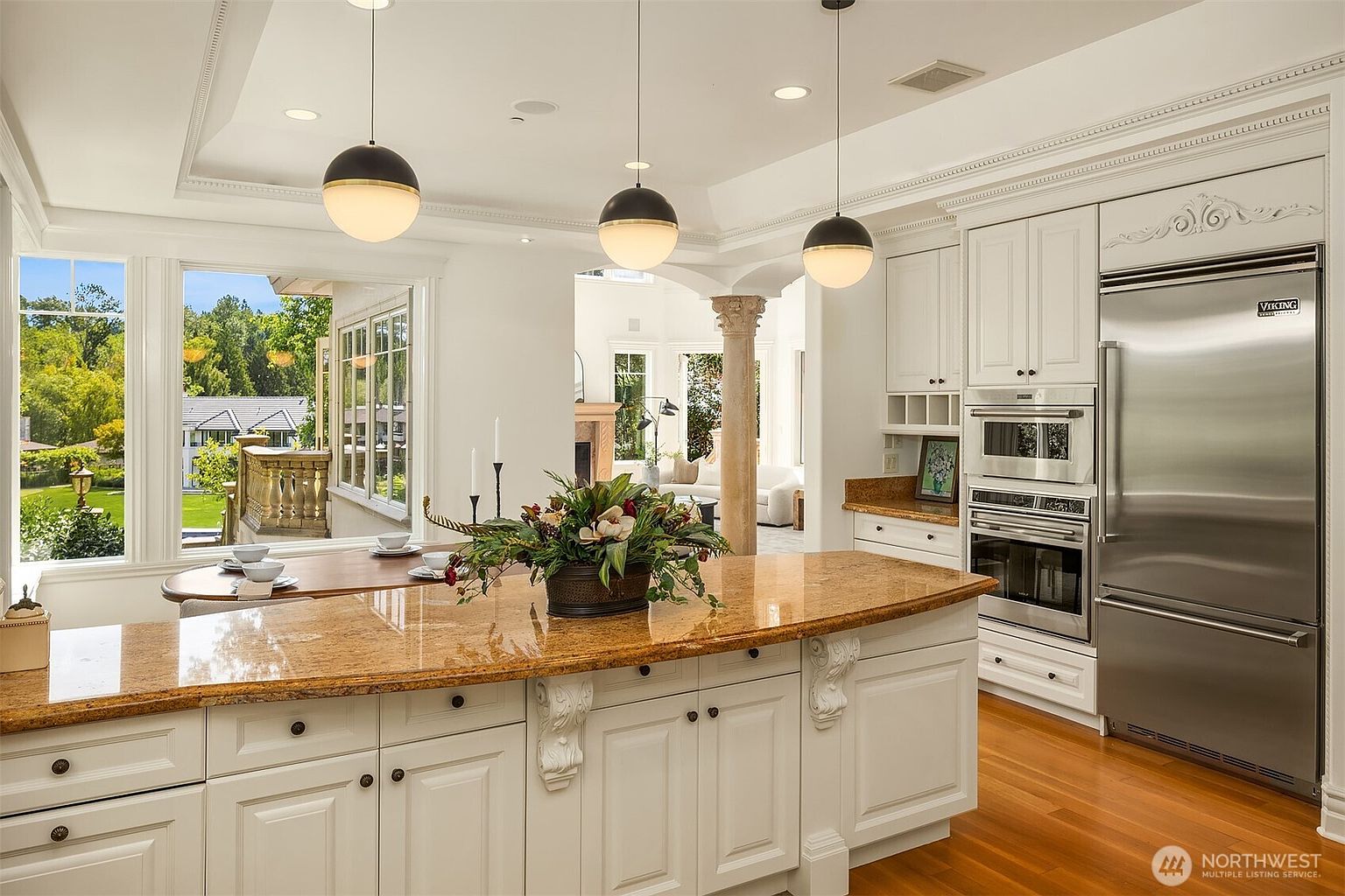 This elegant kitchen features a large granite-topped island with ornate corbel detailing, complemented by white cabinetry and high-end stainless steel Viking appliances. Three modern globe pendant lights hang above the island, while large windows provide a bright, airy view of the lush green backyard. The space seamlessly transitions into an adjacent living area, showcasing a sophisticated and open-concept design.