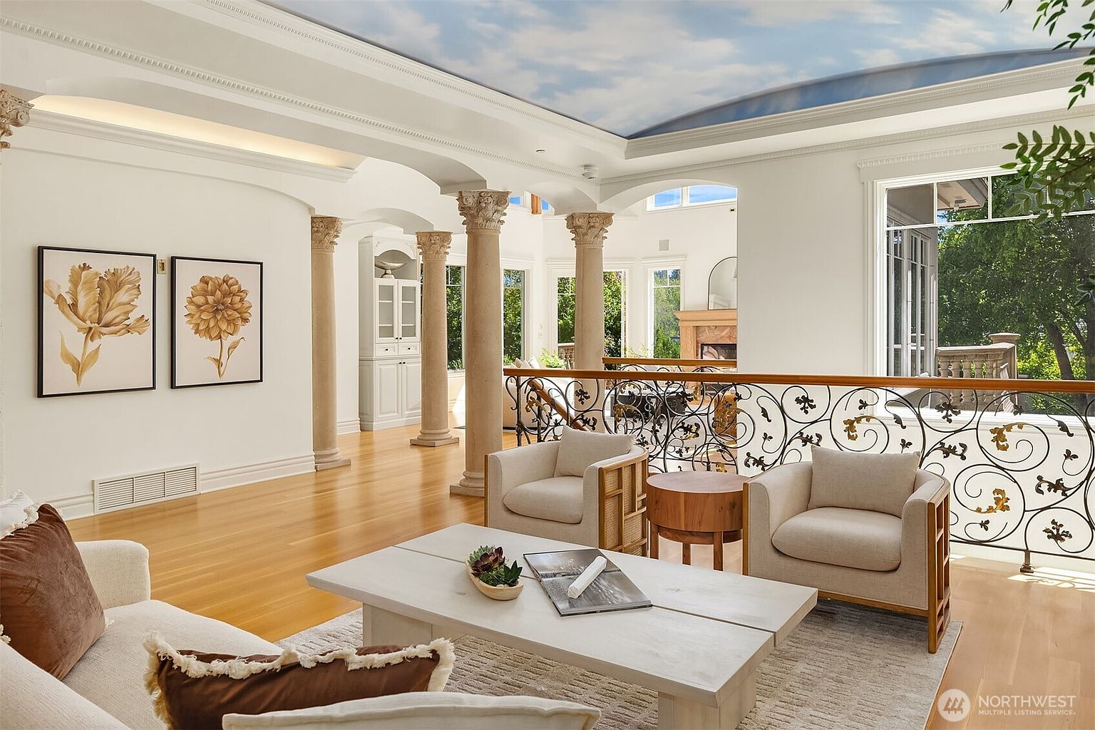 This elegant, open-concept living area features a dramatic sky-painted ceiling and classical columns that create a sophisticated, airy atmosphere. The space is furnished with plush, neutral-toned seating, a rustic wooden coffee table, and intricate wrought-iron railings overlooking a lower level. Large windows flood the room with natural light, highlighting the polished hardwood floors and refined architectural details.
