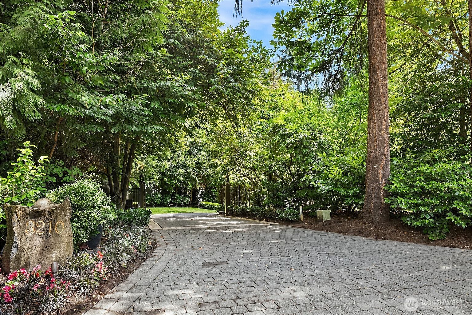 This image captures a private, paved driveway leading into a lush, tree-lined property, marked by a prominent stone address marker reading '3210'. The scene is framed by mature trees and dense greenery, creating a secluded and serene atmosphere. The perspective is from the street level, inviting the viewer to follow the winding path toward the hidden residence.