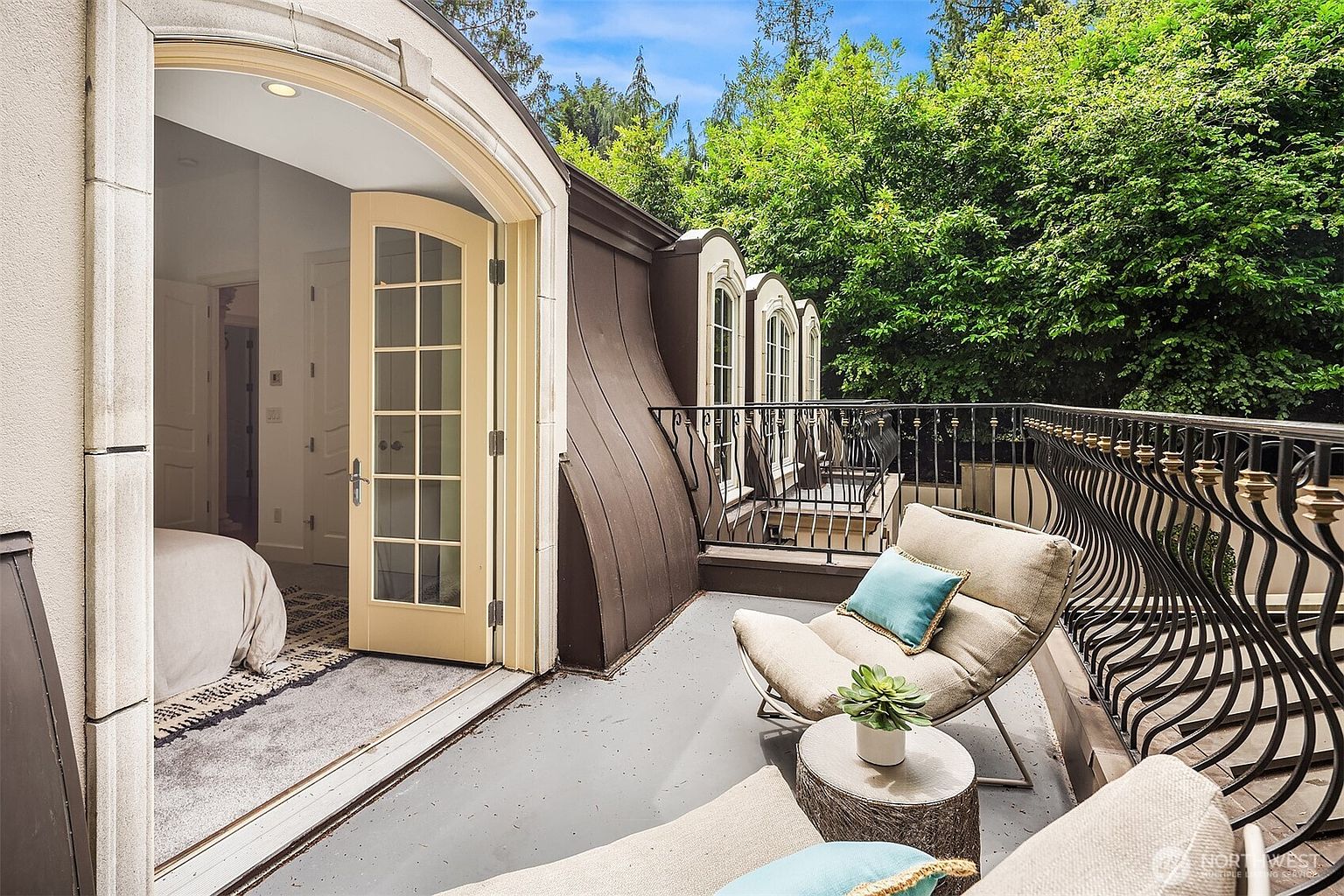 This elegant balcony offers a serene outdoor retreat, featuring a pair of comfortable lounge chairs and a small side table with a potted succulent. The space is framed by a classic wrought-iron railing and overlooks lush, mature trees, while a set of French doors provides seamless access to the adjacent bedroom. The architectural design includes a distinctive curved roofline, creating a sophisticated and inviting atmosphere.