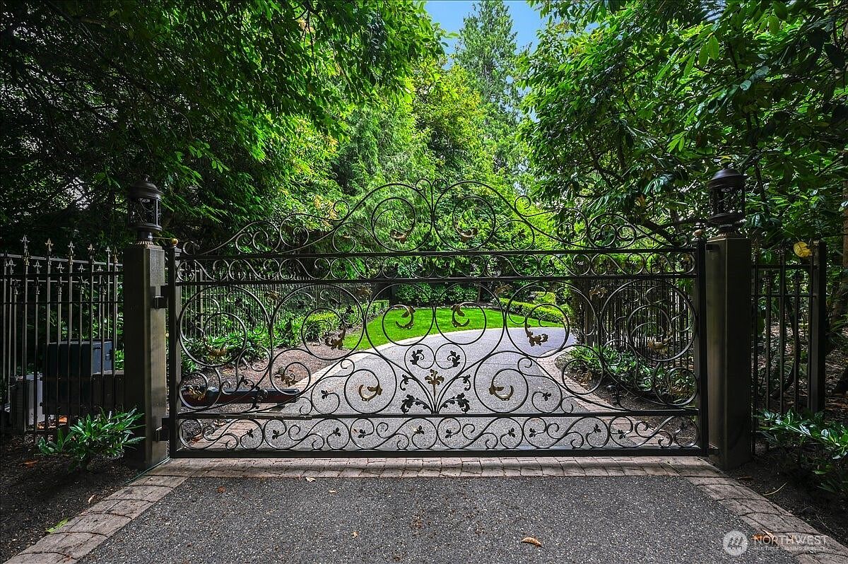 This image captures a grand, ornate wrought-iron gate serving as the primary entrance to a private estate. The intricate metalwork features elegant scroll patterns and decorative accents, framed by lush, mature greenery and trees that create a sense of seclusion and prestige. The perspective is a straight-on, eye-level shot that emphasizes the imposing and sophisticated nature of the property's entrance.