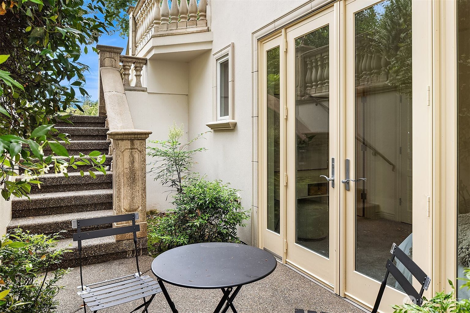 This charming outdoor patio space features a small bistro set placed on a textured concrete surface, adjacent to a set of elegant French doors. The area is framed by a classic stone staircase with ornate balustrades, creating a sophisticated and private atmosphere. The combination of lush greenery and architectural stonework provides a serene, European-inspired retreat perfect for morning coffee.
