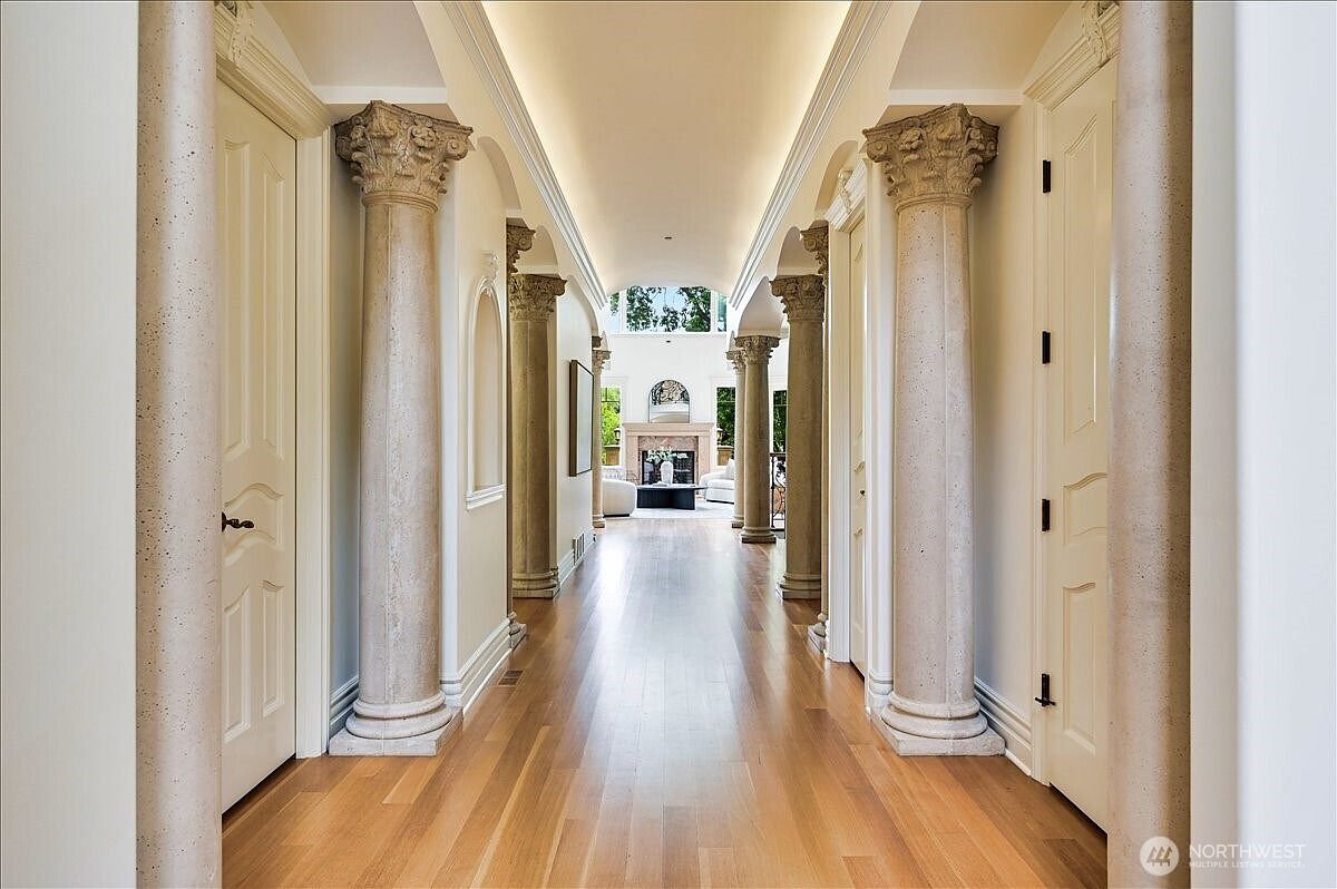 This elegant, grand hallway features a series of classical columns with ornate capitals that line the path, creating a sophisticated and symmetrical architectural aesthetic. The polished hardwood floors lead the eye toward a bright, open living space at the end of the corridor, which is illuminated by natural light from large windows. The high, coved ceiling with recessed lighting enhances the sense of luxury and spaciousness throughout the home.