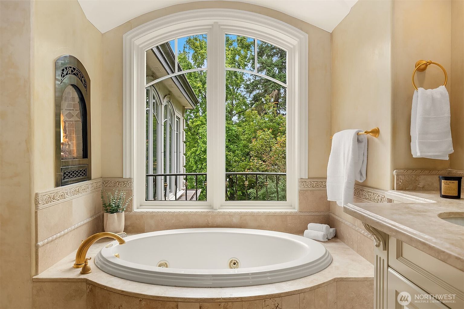 This luxurious primary bathroom features a large, oval jetted soaking tub centered beneath a grand arched window that offers a serene view of the surrounding greenery. The space is finished with elegant beige stone tile, a built-in fireplace to the left, and gold-toned fixtures that add a touch of sophistication. The overall atmosphere is tranquil and spa-like, emphasizing comfort and high-end design.