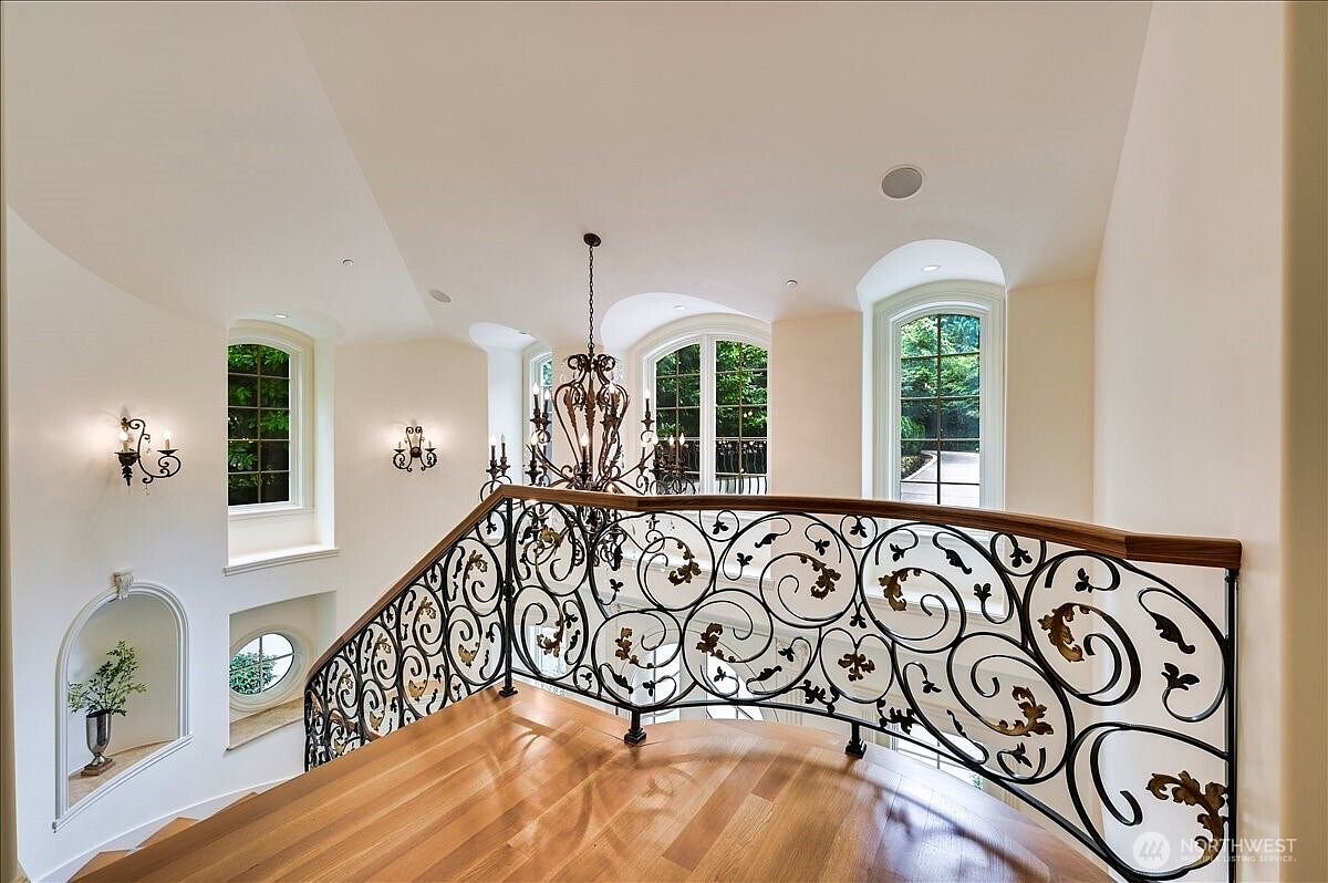 This elegant landing area features a sophisticated wrought-iron railing with ornate scrollwork and a warm wood handrail, overlooking a grand staircase. The space is bathed in natural light from multiple arched windows, highlighting the polished hardwood floors and a decorative wall niche with a potted plant. A large, intricate chandelier serves as a dramatic focal point, enhancing the luxurious and classic architectural style of the home.