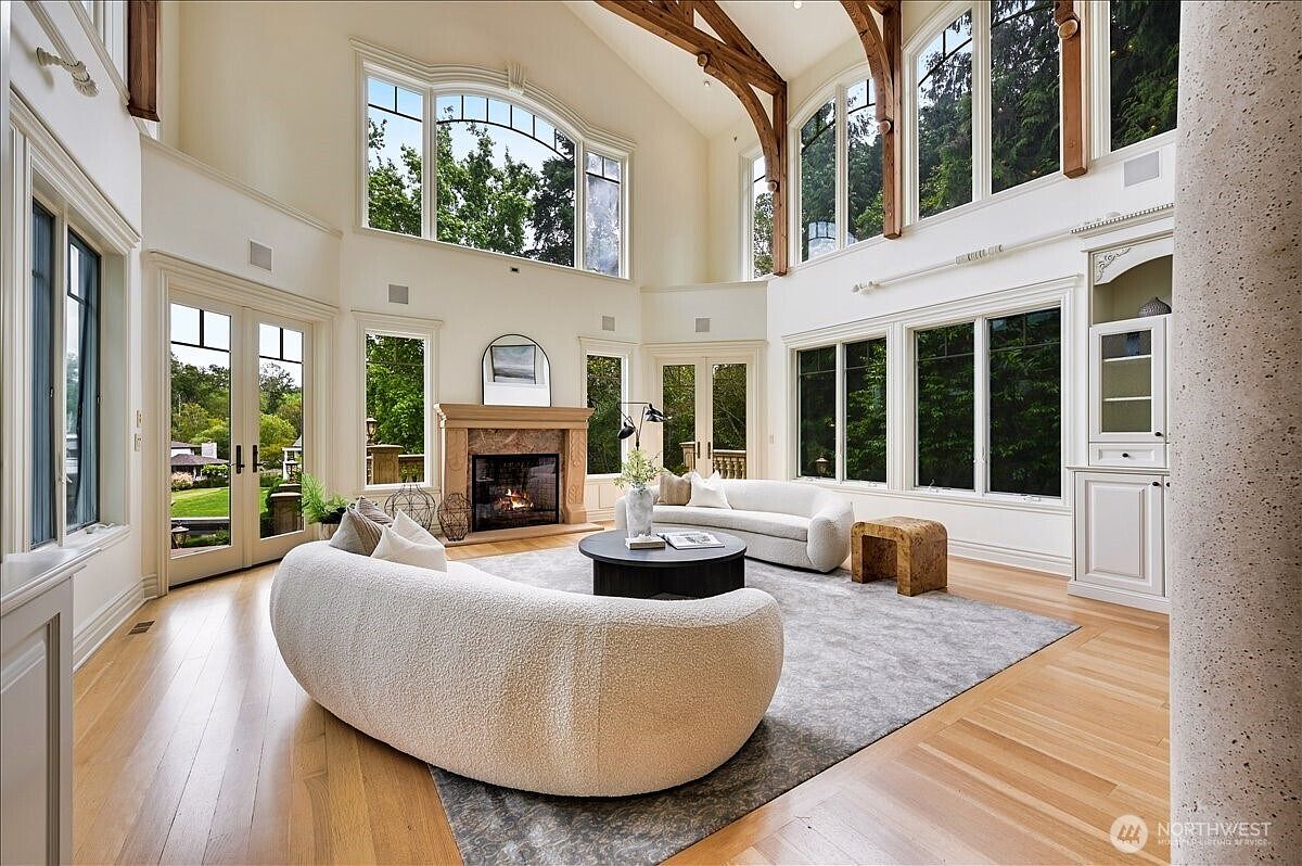 This expansive, light-filled living room features soaring vaulted ceilings with exposed wooden beams and massive floor-to-ceiling windows that offer a panoramic view of the surrounding greenery. The space is anchored by a grand stone fireplace and furnished with elegant, curved white sofas arranged around a central dark circular coffee table on a large area rug. The room exudes a sophisticated, airy, and luxurious atmosphere, perfect for high-end entertaining.
