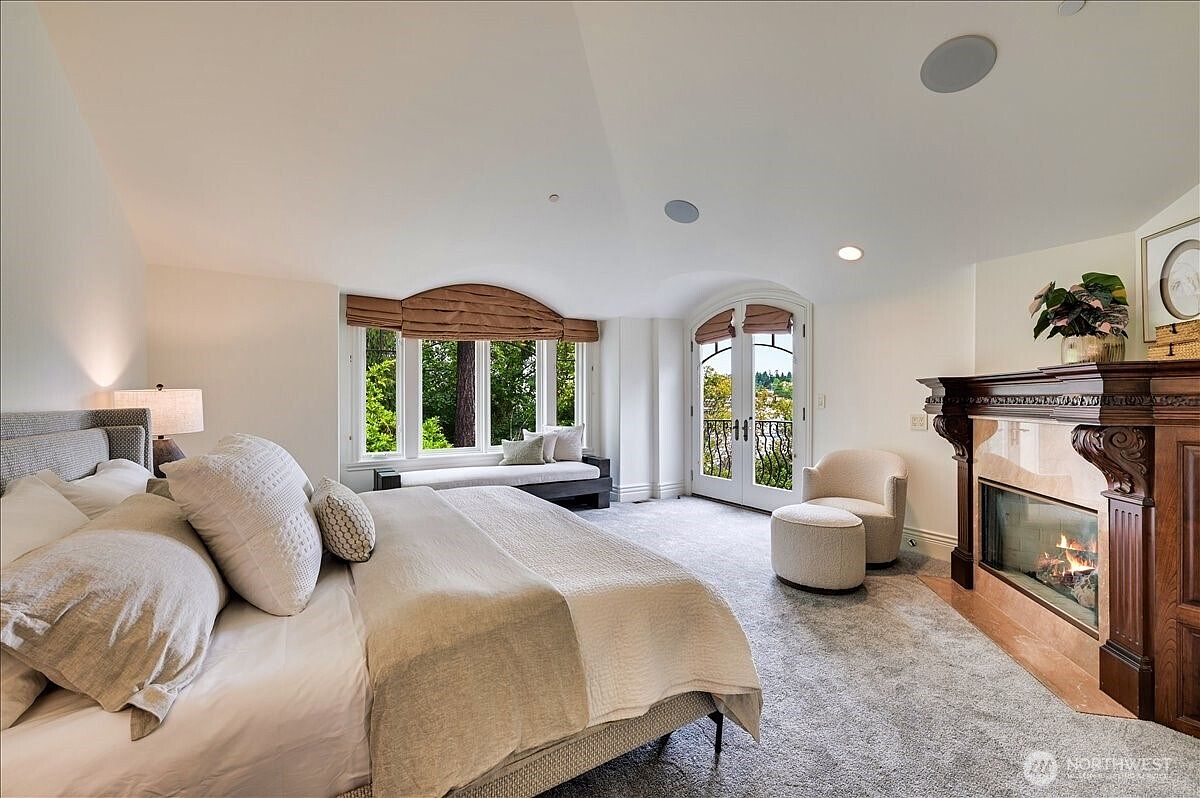 This elegant primary bedroom features a plush king-sized bed, a cozy window seat with custom drapery, and a classic fireplace with a dark wood mantel. The room is bathed in natural light from the large windows and French doors, which open to a view of the surrounding greenery. The neutral color palette and soft carpeting create a serene and luxurious atmosphere.