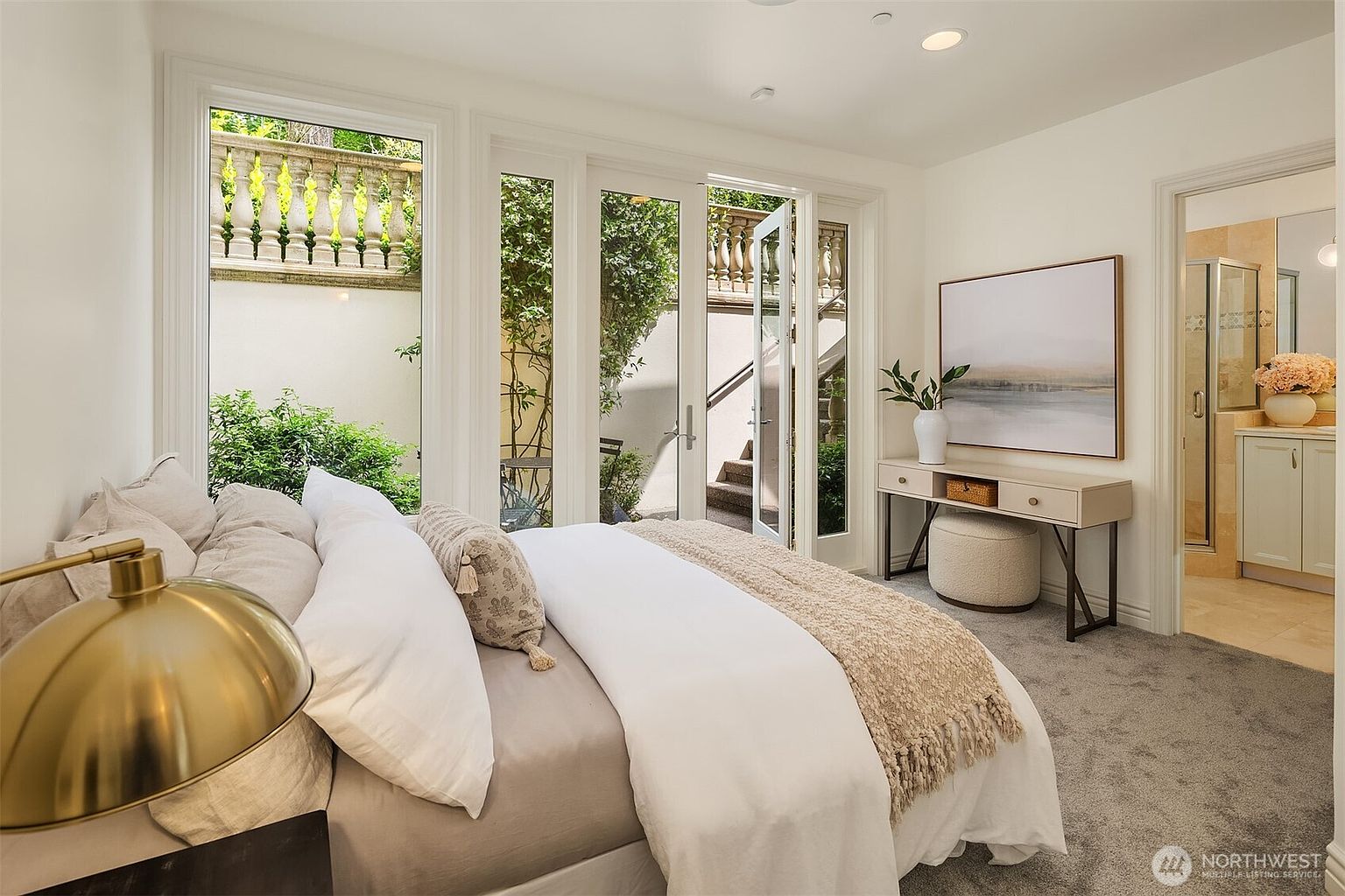 This bright and airy primary bedroom features a plush bed with neutral-toned bedding, positioned next to floor-to-ceiling windows and glass doors that open to a private, landscaped courtyard. A sleek console table with a decorative vase and artwork adds a touch of elegance, while the adjacent doorway provides a glimpse into a well-appointed bathroom. The room exudes a serene and sophisticated atmosphere, enhanced by soft carpeting and clean, minimalist decor.