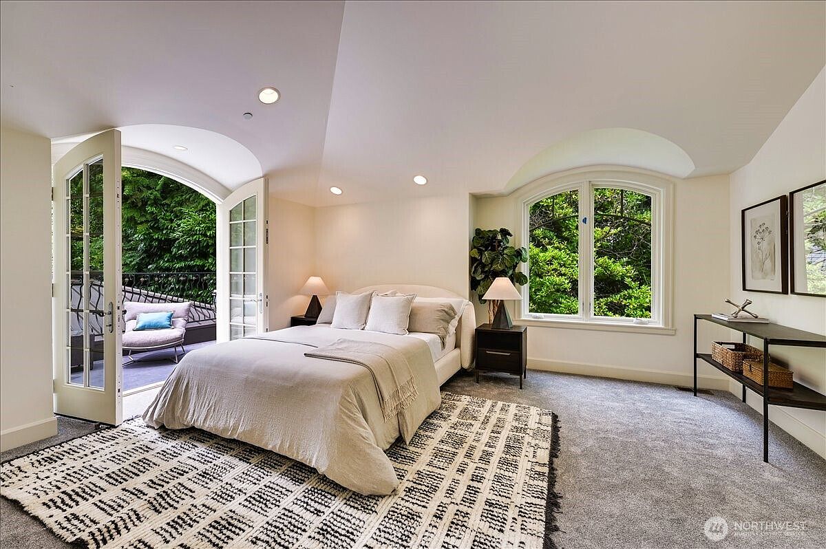 This bright and airy primary bedroom features a neutral color palette, plush carpeting, and a large bed centered in the room. The space is highlighted by elegant arched French doors that open onto a private balcony, allowing for abundant natural light and a seamless indoor-outdoor connection. A sleek console table and minimalist decor contribute to a sophisticated, tranquil atmosphere.