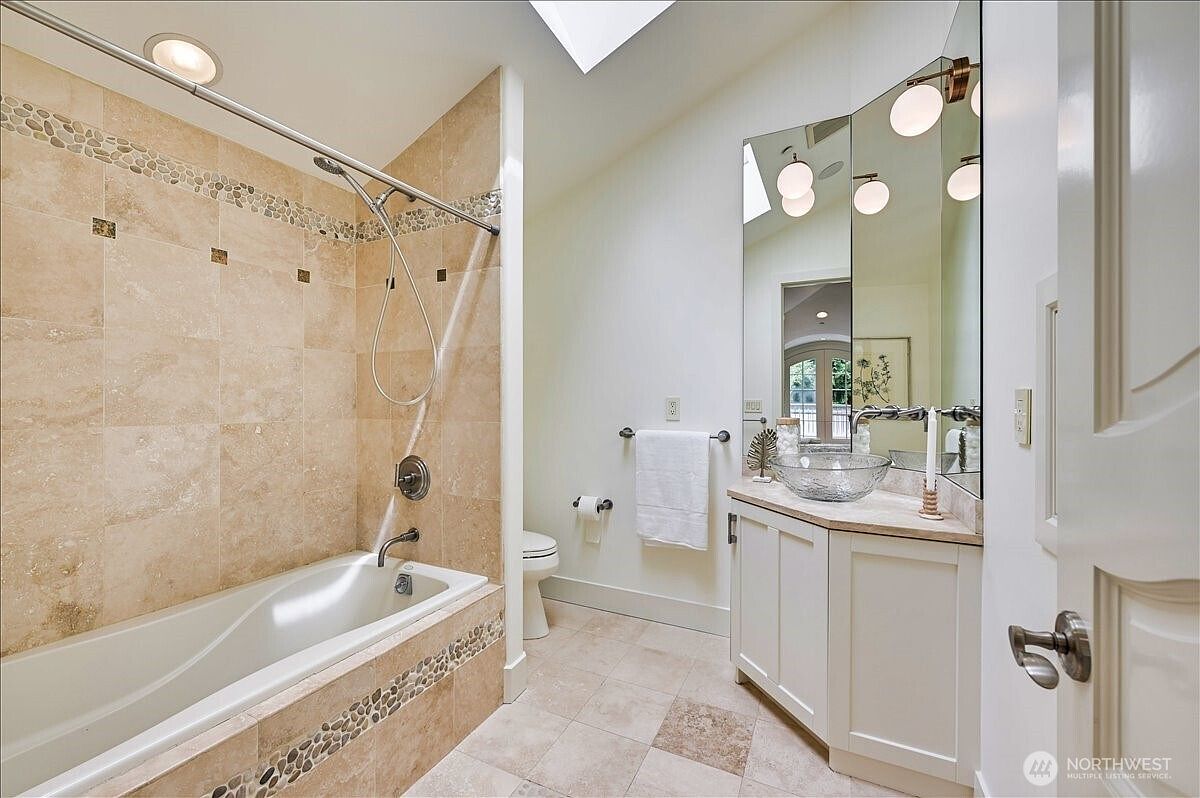 This bright and airy bathroom features a combination tub and shower with neutral stone tile surround and a decorative mosaic accent band. The vanity is equipped with a unique glass vessel sink and a large, multi-paneled mirror that reflects the natural light from the overhead skylight. The space is finished with light-colored cabinetry and tile flooring, creating a clean and inviting atmosphere.