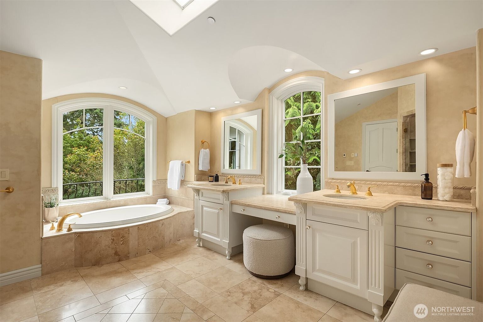 This luxurious primary bathroom features a sophisticated design with dual vanities, a central makeup station, and a deep soaking tub nestled beneath a large arched window. The space is finished with elegant cream-toned cabinetry, marble countertops, and light-colored tile flooring, creating a bright and serene atmosphere. The high, vaulted ceiling and ample natural light enhance the sense of openness and refinement in this spa-like retreat.