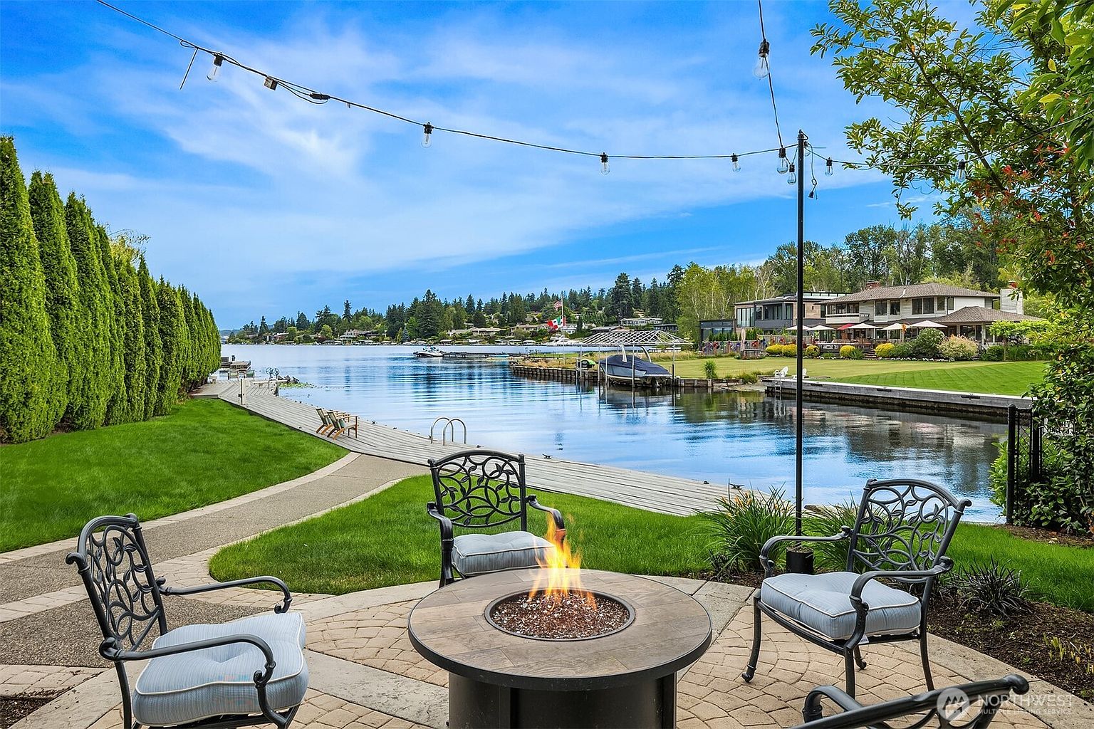 This serene waterfront patio features a circular fire pit surrounded by elegant wrought-iron chairs, creating an inviting outdoor living space. The scene overlooks a calm lake with a private wooden dock, lush green lawns, and a neighboring luxury home in the distance. String lights overhead add a warm, cinematic touch to this picturesque lakeside setting.