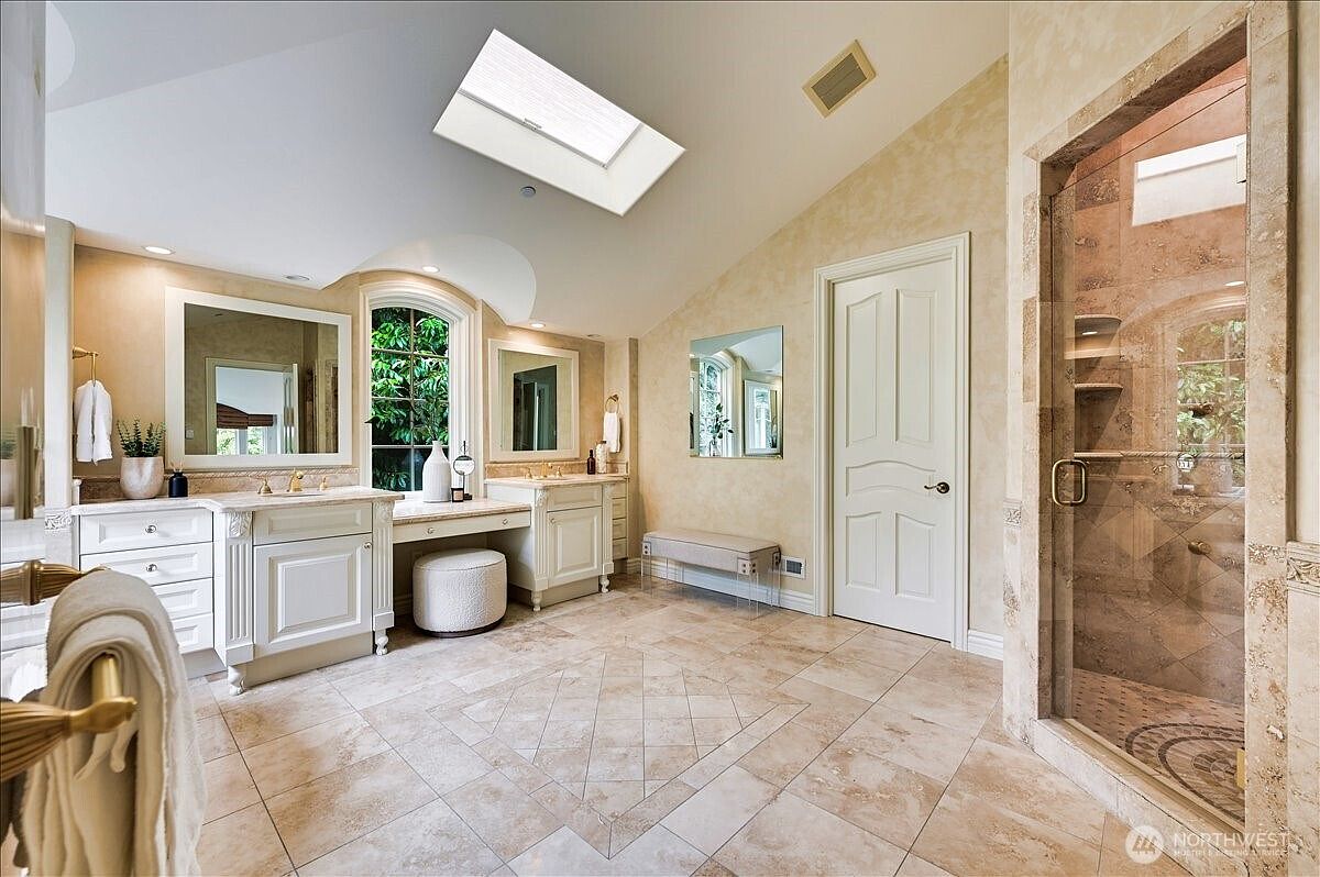 This luxurious primary bathroom features a dual-vanity setup with elegant white cabinetry, marble countertops, and a central makeup vanity area. The space is brightened by a large skylight and a tall arched window, while a spacious walk-in glass shower with stone tile finishes adds a sophisticated touch. The overall aesthetic is refined and airy, with neutral tones and high-end finishes creating a spa-like atmosphere.