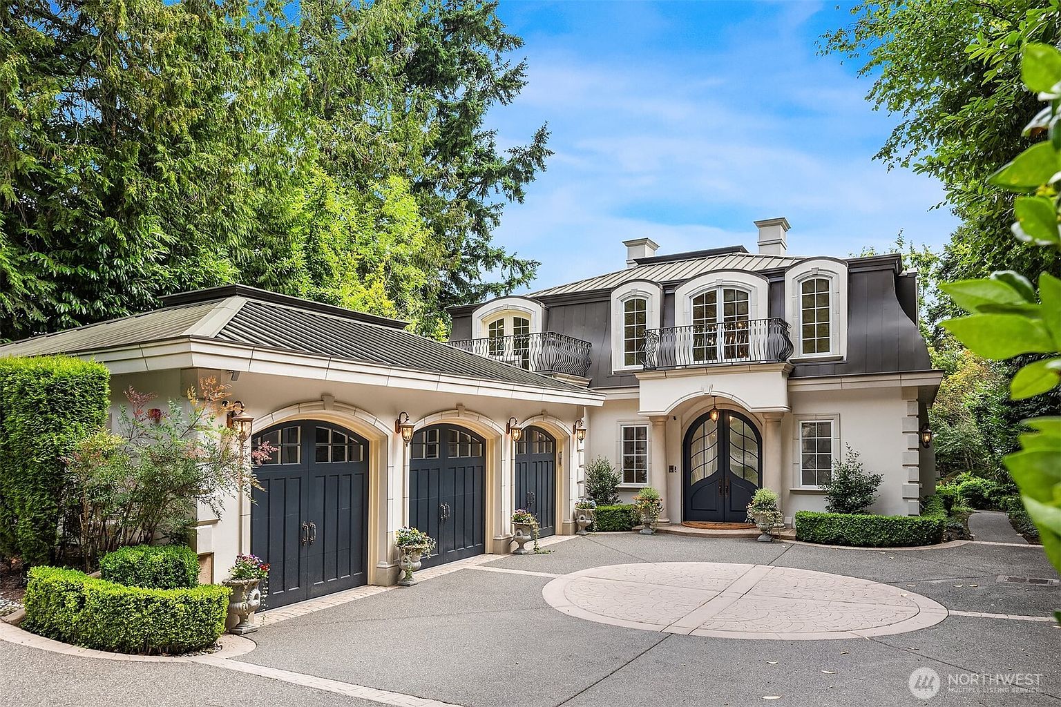 This elegant French-inspired estate features a striking facade with a mansard roof, arched windows, and a prominent balcony above the grand arched entryway. The property includes a spacious three-car garage with matching dark-toned doors and a meticulously landscaped circular driveway. The overall impression is one of sophisticated luxury, set against a backdrop of lush, mature trees.