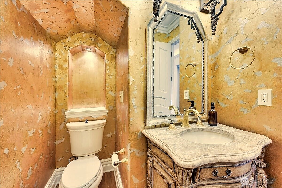 This elegant powder room features a dramatic gold-toned faux finish on the walls, creating a luxurious and warm atmosphere. The space includes a classic pedestal-style vanity with a marble countertop, an ornate framed mirror, and a toilet tucked into a recessed alcove. The overall aesthetic is opulent and traditional, highlighted by gold-toned hardware and sophisticated lighting fixtures.